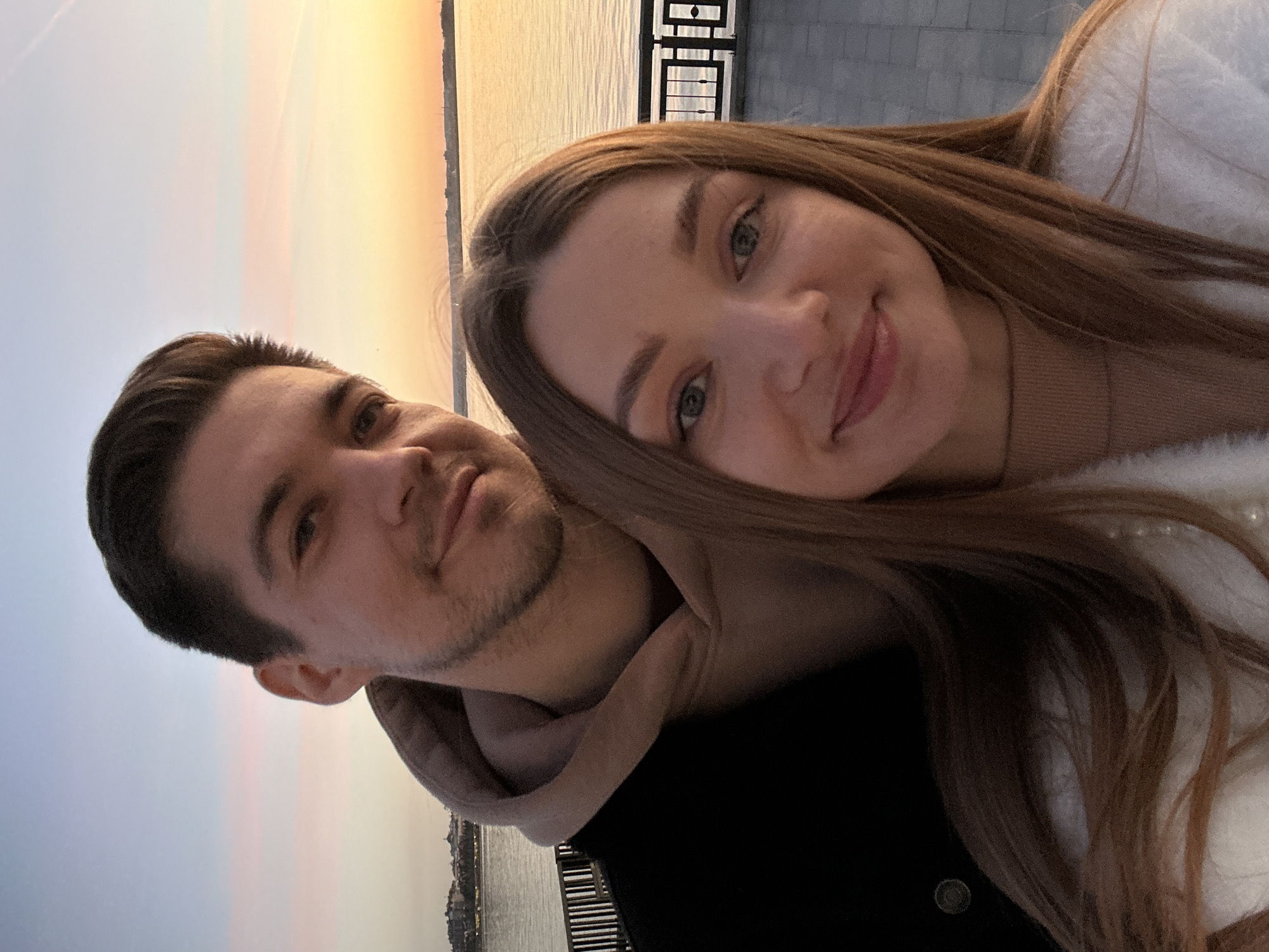A close-up portrait-oriented image features a man and a woman together, likely a selfie taken against a picturesque waterfront backdrop during sunset or sunrise in Kazan, Russia. The image, originally rotated, shows the man positioned on the left and the woman on the right, with her head resting on his shoulder.
The man has dark hair and light stubble or a short beard. He is wearing a light-colored hoodie under a dark outer garment, and he looks towards the camera with a subtle smile. The woman has long, straight brown hair and blue eyes, and she is smiling warmly at the camera. She wears a light-colored, possibly fuzzy, sweater or top over a light brown base layer.
Behind the couple, the sky showcases a vibrant sunset or sunrise with a soft gradient of colors, ranging from light blue at the top to warm oranges, yellows, and pinks near the horizon. Below the colorful sky lies a calm body of water, presumably the Volga River or another body of water in Kazan, with a faint cityscape visible on the distant bank. A dark metal railing with geometric designs runs along what appears to be an embankment or promenade, adding a structural element to the scene.
Both individuals exude happiness and affection, creating an intimate and pleasant atmosphere. The clear sky and beautiful natural light indicate fair weather conditions. No visible text is present in the image.