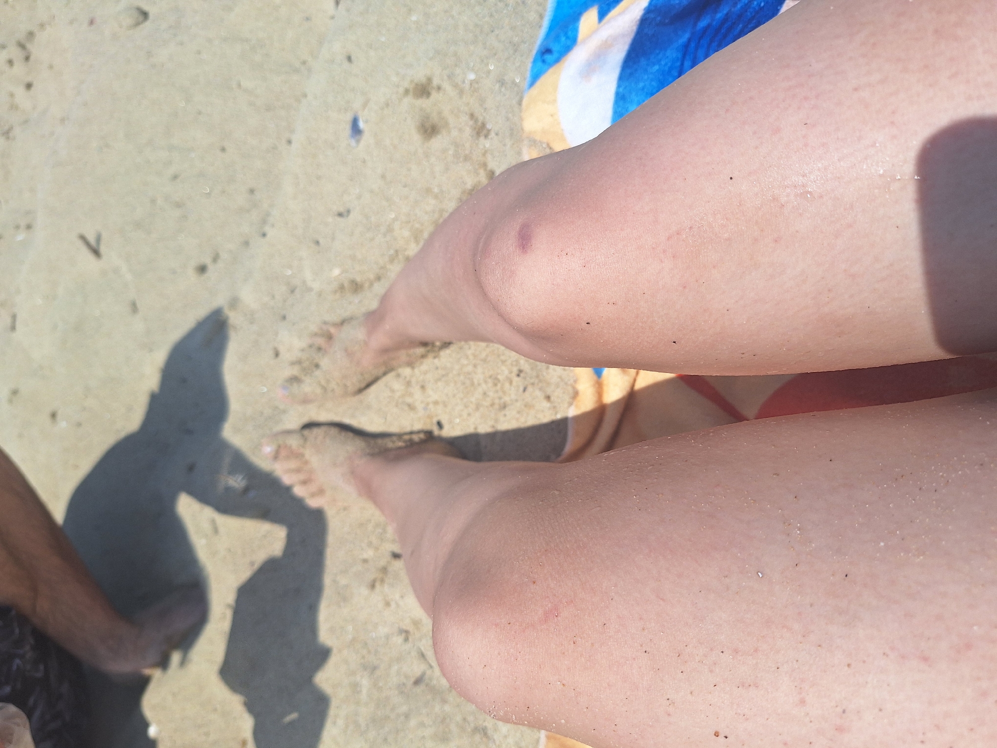 The image shows a close-up view of two pairs of legs on a sandy beach. The foreground is dominated by the legs of a person lying down, with sand visible on their skin and knees. A beach towel with blue and white stripes is partially visible beneath the legs. In the background, the legs and feet of another person are visible, as well as shadows cast on the sand. The setting is a beach, likely in Bliznatsi, Bulgaria, given the provided location context. The overall scene depicts a casual moment of relaxation on the beach.