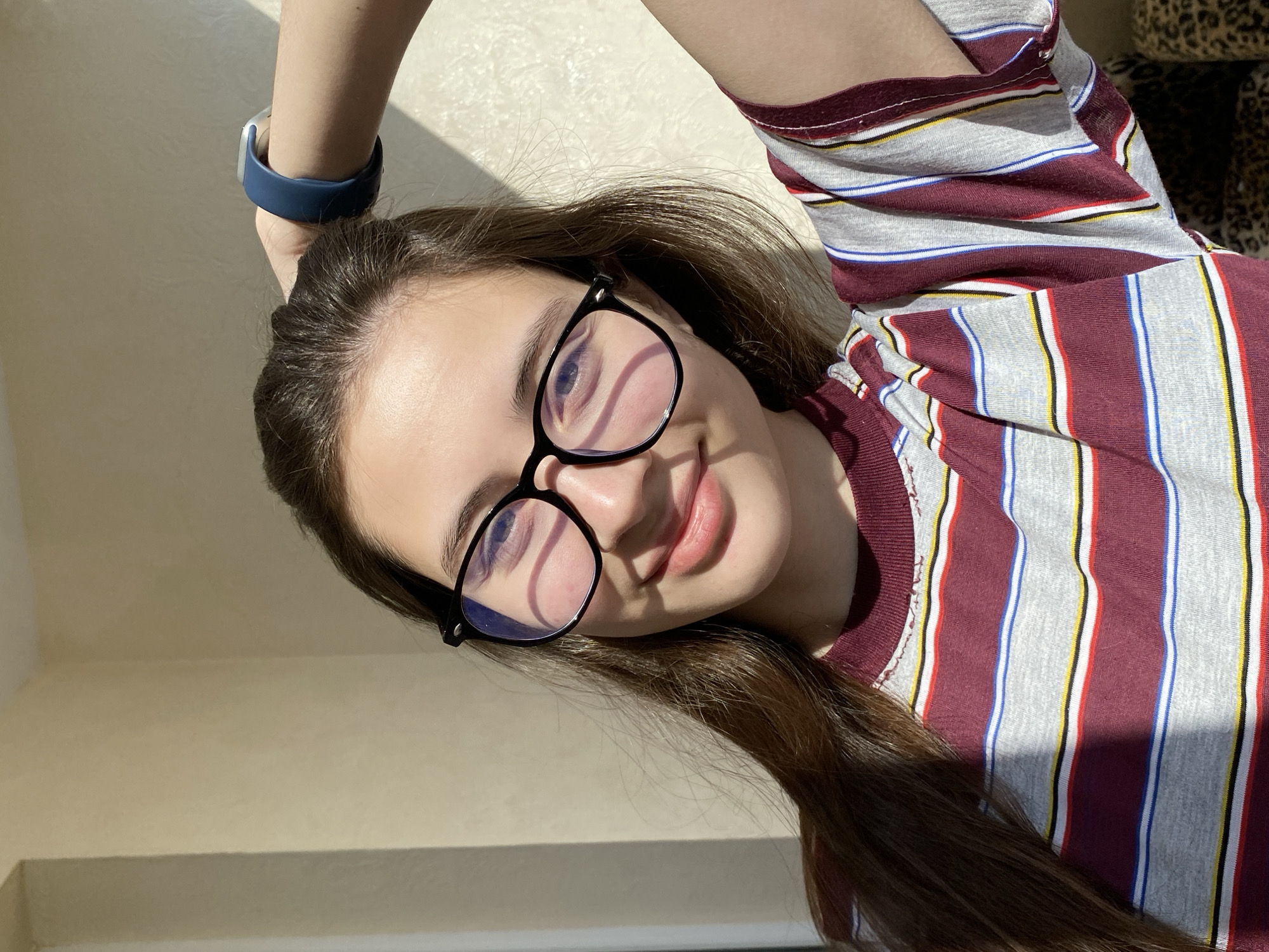 A close-up, slightly tilted portrait features a young woman indoors in Votkinsk, Russia. She has long, dark brown hair and is wearing black-rimmed glasses, which show reflections of the light source. A gentle smile graces her lips, and her gaze is directed towards the camera.
She is dressed in a casual striped t-shirt with vertical bands of maroon, light grey, white, blue, red, and yellow. On her left wrist, a blue-banded smart device or watch with a silver component is visible. Her left arm is raised, with only the forearm and wrist visible in the frame.
The setting is an indoor space with light-colored, plain walls, bathed in strong, directional sunlight that creates distinct shadows, indicating it is daytime. A hint of a patterned surface, possibly leopard print, can be seen in the upper right corner of the frame, suggesting furniture or a fabric item nearby. The woman's expression conveys a calm and pleasant mood. No visible text is present in the image.