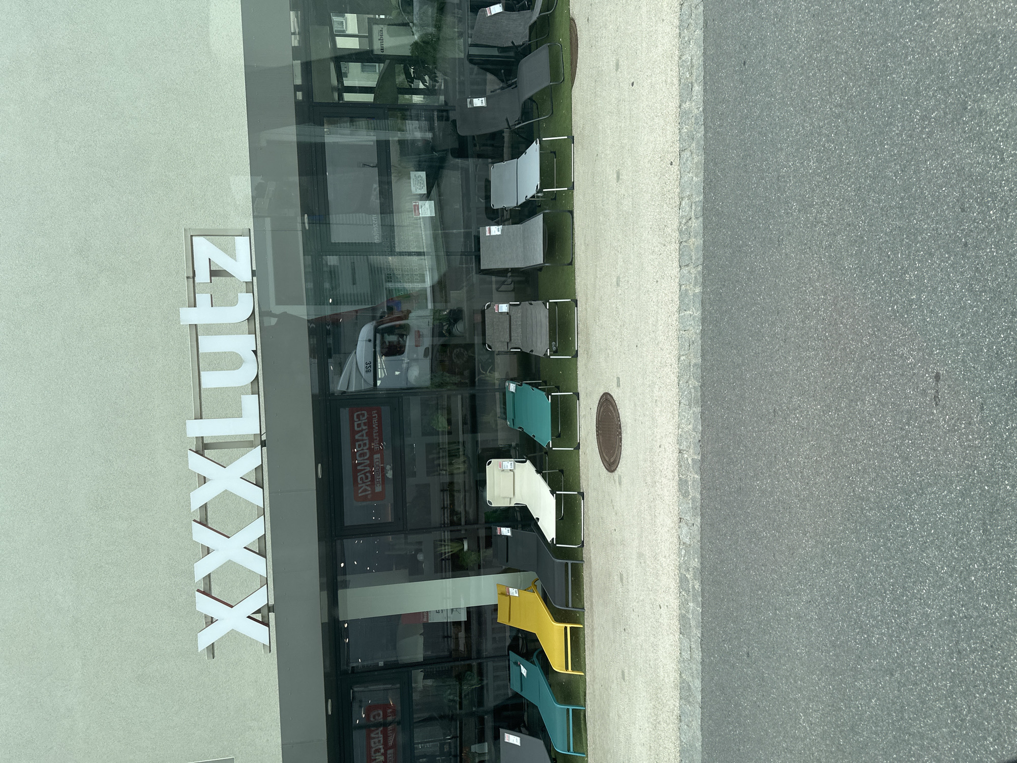 The image shows the XXXLutz furniture store located in Lauterach, Austria. The storefront features large glass windows displaying a row of outdoor lounge chairs on a green artificial turf surface. The chairs are in various colors, including teal, yellow, dark gray, and cream. A manhole cover is visible on the pavement in front of the store. Reflections of a truck and other elements are visible in the glass windows. The XXXLutz logo is prominently displayed on the building's facade.