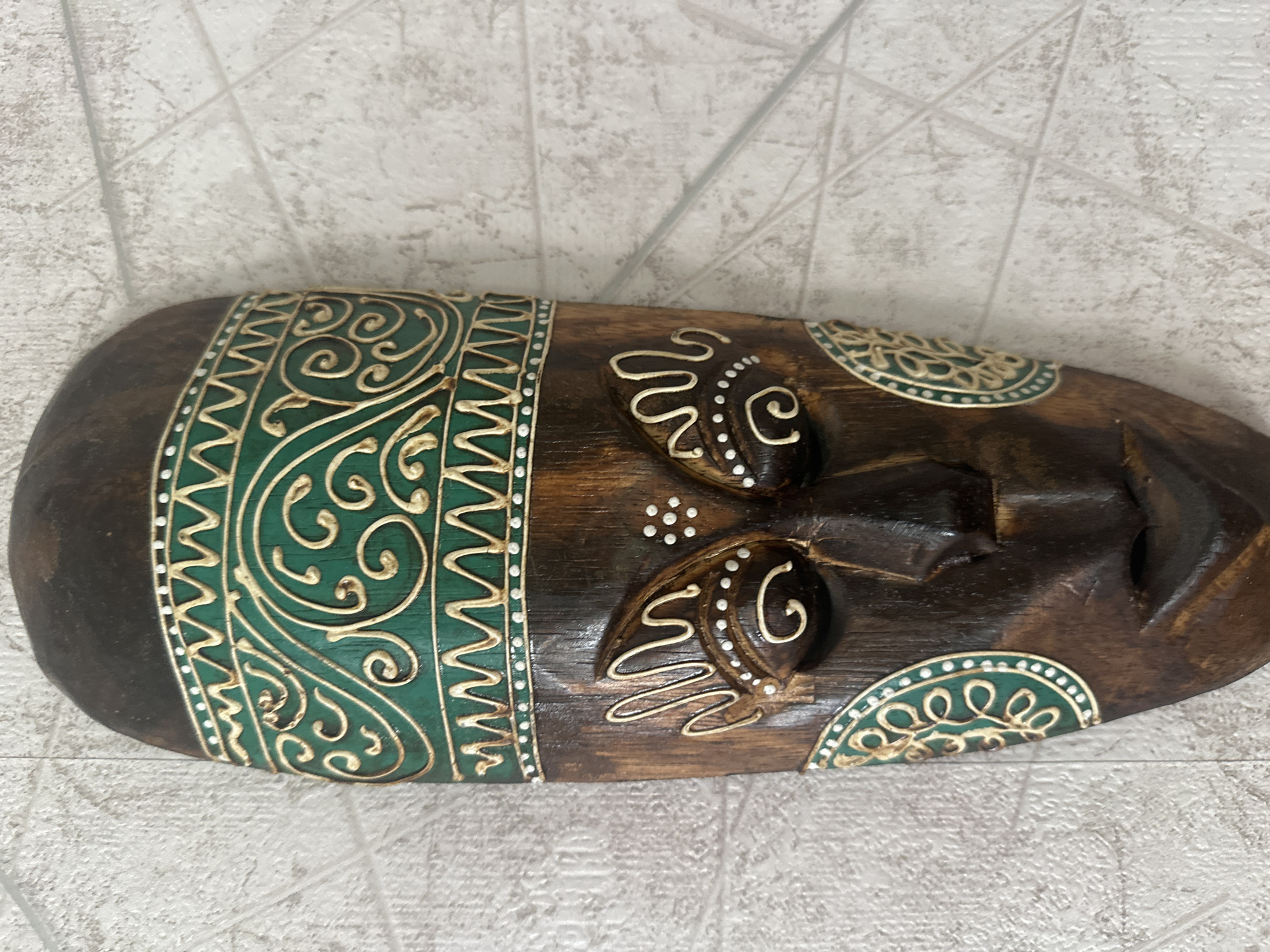 This is an image of a decorative wooden mask, likely intended as a wall hanging or cultural artifact. The mask is dark brown wood with intricate carvings and painted details. The left side of the mask features a wide band with a geometric pattern and swirling green and gold decorative elements. The central part of the mask depicts facial features, including raised eye areas adorned with white swirling lines and white dots. The nose is carved and prominent, and there is an opening for the mouth. The right side of the mask shows a partial circular decorative element, also in green and gold, with a leaf-like pattern. The mask is lying horizontally on a textured surface that resembles a light-colored tiled wall. The lighting appears to be even, suggesting an indoor setting. There is no visible text in the image. The mask itself suggests a handcrafted item with a tribal or ethnic aesthetic, possibly inspired by African or other cultural art forms.