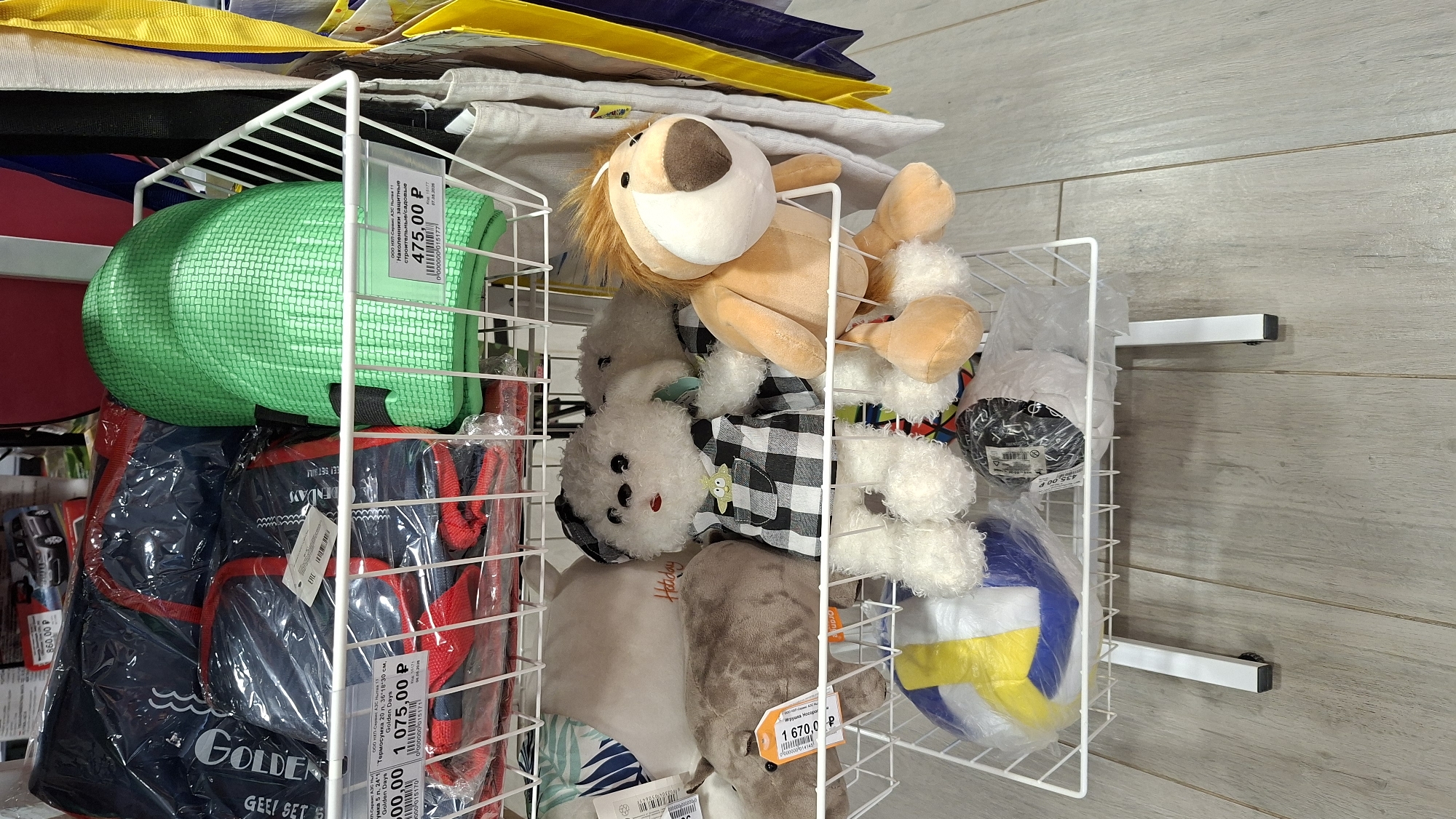 The image displays the interior of a retail store in Nytva, Russia, showcasing various items arranged on white wire shelving units. The foreground features a white wire rack holding several plush toys, including a white dog wearing a checkered outfit, a light brown lion, and a gray hippopotamus. A colorful volleyball is also visible in the lower section of the rack. Behind the plush toys, on a higher shelf, are green knee pads and a blue and red sports bag. The background reveals more merchandise and store fixtures, including yellow and blue shopping bags hanging on the right. The flooring appears to be light-colored wood laminate. The scene is static and depicts a typical retail display.