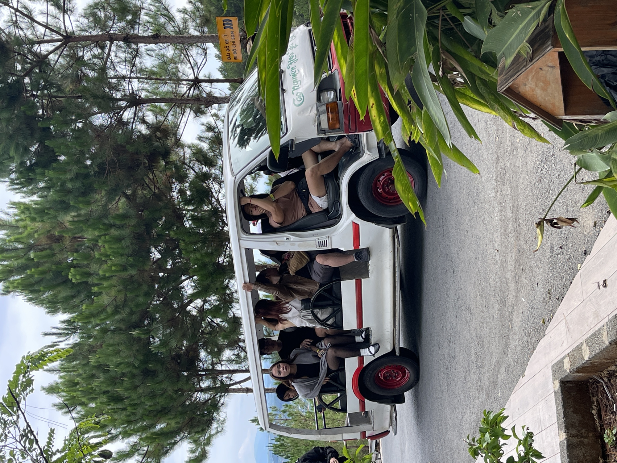 A white bus-like vehicle with an open-air passenger area is parked on a paved road, oriented at an angle in the frame. The location is identified as Thôn 5, Vietnam by the presence of a sign in Vietnamese. People are seated within the vehicle’s open passenger area. The vehicle is parked near a tree-lined area. The road surface appears to be asphalt or concrete. Tropical vegetation is present in the foreground. A sign is visible at the top right of the frame.