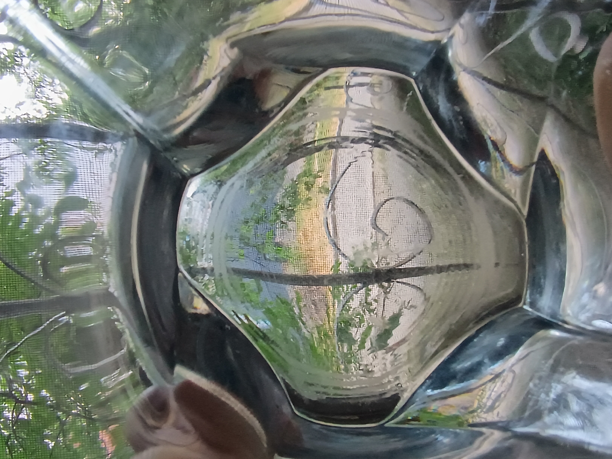 The image shows a close-up, distorted reflection of trees and a screened window on the curved surface of a glass or plastic object. The reflection is warped and fragmented, with some areas appearing brighter or darker than others. A faint heart shape is visible in the reflection, possibly etched or drawn on the screen. The object itself appears to be clear with a textured or faceted surface, creating the distortion. The background suggests an outdoor environment with greenery and a structure behind the screen, but these details are heavily obscured by the reflection and distortion. The image does not provide any information or visual cues that would allow for the identification of its location within the city of Zaporizhzhia, Ukraine.