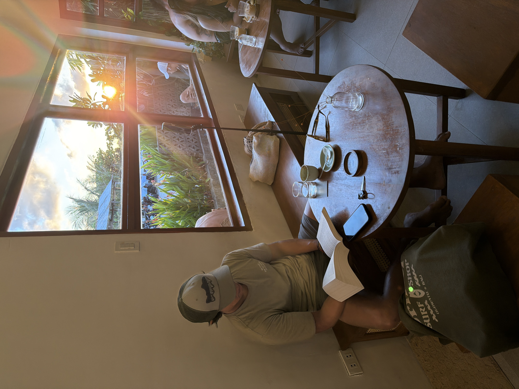A person wearing a baseball cap and a t-shirt sits and reads a book indoors. A phone rests on the table to the right. A table is set with cups, saucers, and a glass bottle. Outside the window, trees are visible. The location appears to be a cafe or similar establishment in General Luna, Philippines.