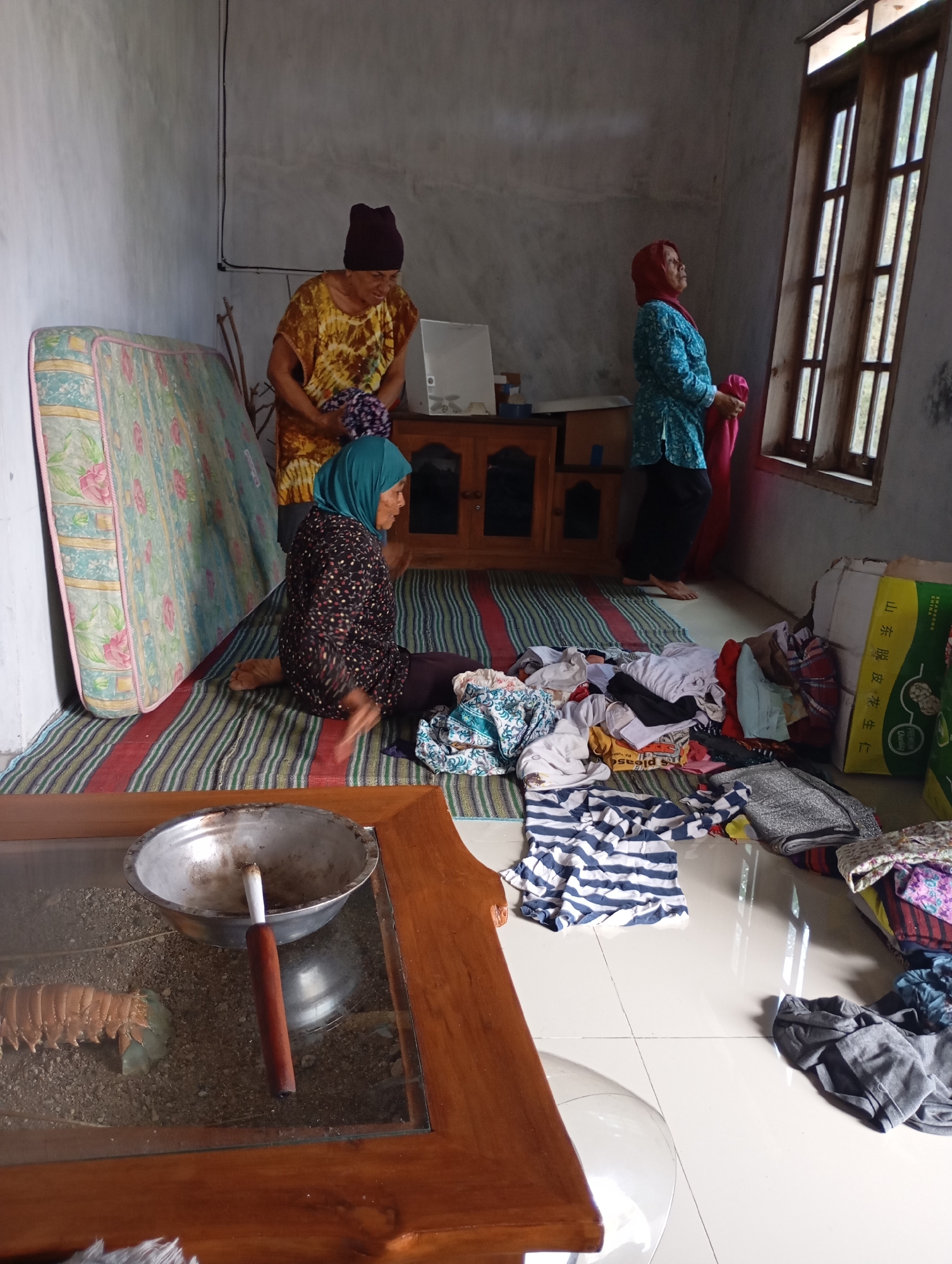 In a room that appears to be in Ketepung, Indonesia, three women are engaged in activities related to laundry or clothing. One woman, wearing a maroon headscarf and a yellow and brown patterned top, stands near a mattress and a wooden cabinet, holding a folded piece of fabric. Another woman, in a blue patterned top and red headscarf, stands by a tall window, also holding a red garment. The third woman, seated on the floor in a black and white patterned top and blue headscarf, is surrounded by a pile of laundry and appears to be sorting or folding clothes. The floor is tiled, and a striped mat is laid out. In the foreground, a wooden table with a glass top holds a metal bowl and a wooden utensil. Various items of clothing are strewn across the floor and on the mat. The room has a rustic and lived-in appearance.