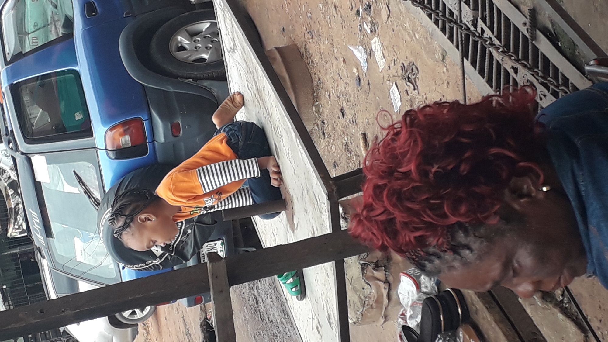 In Onitsha, Nigeria, a young child with braided hair, wearing an orange long-sleeved shirt with white stripes and blue jeans, is sitting on a raised platform. The child's bare feet are visible. To the left of the child, a pair of green sandals rests on the ground. Behind the child, a blue SUV is parked, with a spare tire mounted on its rear. In the foreground, a woman with vibrant red curly hair and a blue denim jacket is partially visible, her head turned to the side. The environment appears to be an outdoor, possibly dusty or muddy area, with discarded items scattered on the ground, including papers and what looks like a metal grate or drain cover. The overall scene depicts a moment in a busy urban setting.