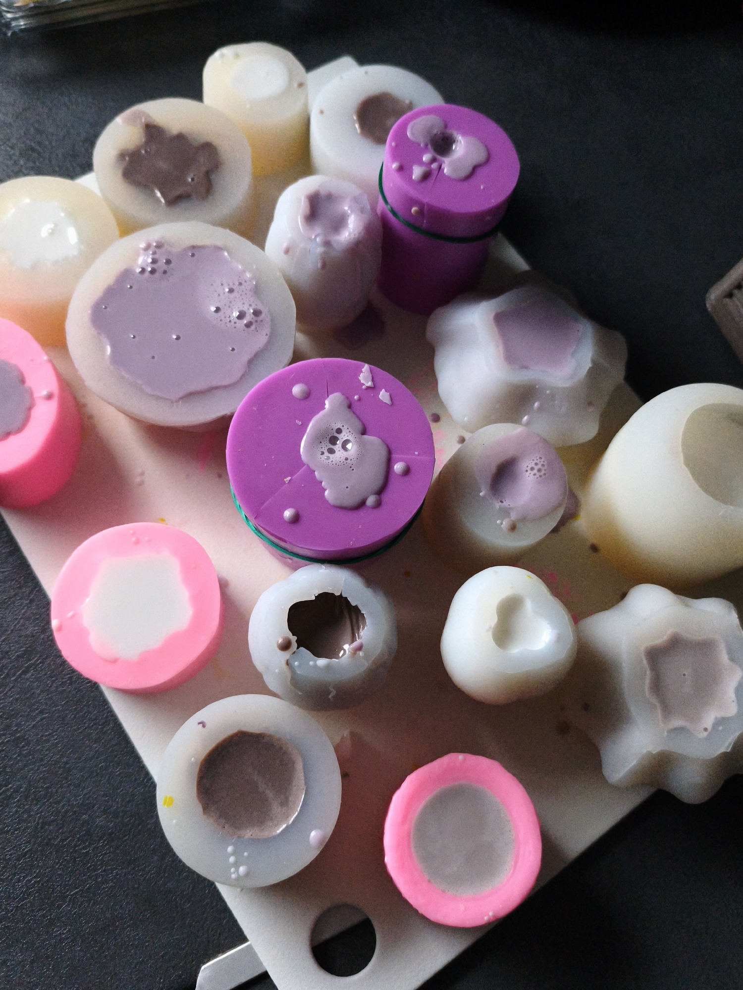 The image displays an overhead view of numerous silicone molds filled with a light purple liquid and some partially dried, darker brown material. The molds are of various shapes and sizes, including circular, cylindrical, and star-like forms. Some molds are completely filled, while others contain only a small amount of liquid or partially cured material. The scene is set on a light-colored, textured surface, likely a mat, with a dark gray background. There are no people visible in the image. The overall impression is of a craft or DIY project in progress, possibly related to soap or candle making. There is no indication of the location being in Brodi, Latvia.
