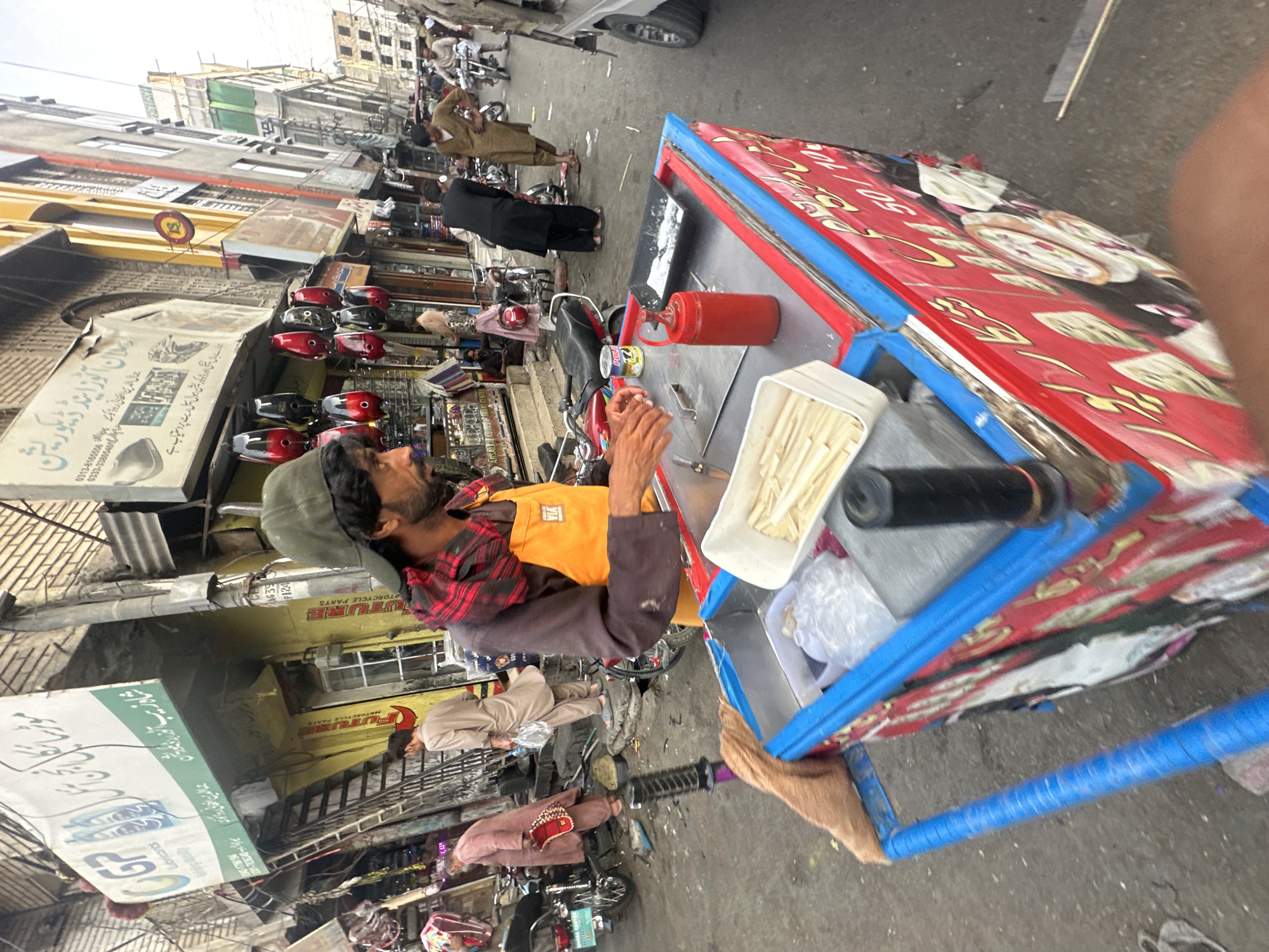 The image captures a vibrant street scene within Quetta City Tehsil, Pakistan. In the immediate foreground, a man with a beard, wearing a green baseball cap, a dark long-sleeved shirt, a red and black plaid scarf, and a bright orange apron with the "VIA" logo, stands behind a red and blue street food cart. His hands are positioned over the cart's metal surface, appearing to prepare an item. On the cart, a white rectangular container holds numerous thin, light-colored sticks (possibly pre-cut food items), alongside a red squeeze bottle, a black tray containing a white powdery substance, and a black cylindrical object. The cart's side features red paneling with prominent yellow Urdu text and images of food items.
The street is paved, exhibiting scattered debris, and is populated by several individuals and vehicles. To the left, a man in a reddish-brown shalwar kameez holds a patterned pouch while standing near a parked red motorcycle. Further back, another man in a light brown shalwar kameez carries a plastic bag. The street is lined with multi-story buildings of varying architectural styles. A large sign on the left building displays "GP Lubricants" and Urdu script, along with "FUTURE MOTORCYCLE PARTS" in yellow lettering below. Hanging from a shop awning on the left are multiple red and black helmet-shaped objects or light fixtures. On the right side of the street, another building features a sign with "ARMAN DECOR" (partially visible) and an orange-striped facade further down. Several men in traditional Pakistani attire are visible walking or riding motorcycles in the mid-ground and background, and a white utility vehicle is partially visible on the far right. The overall environment suggests a busy commercial thoroughfare.