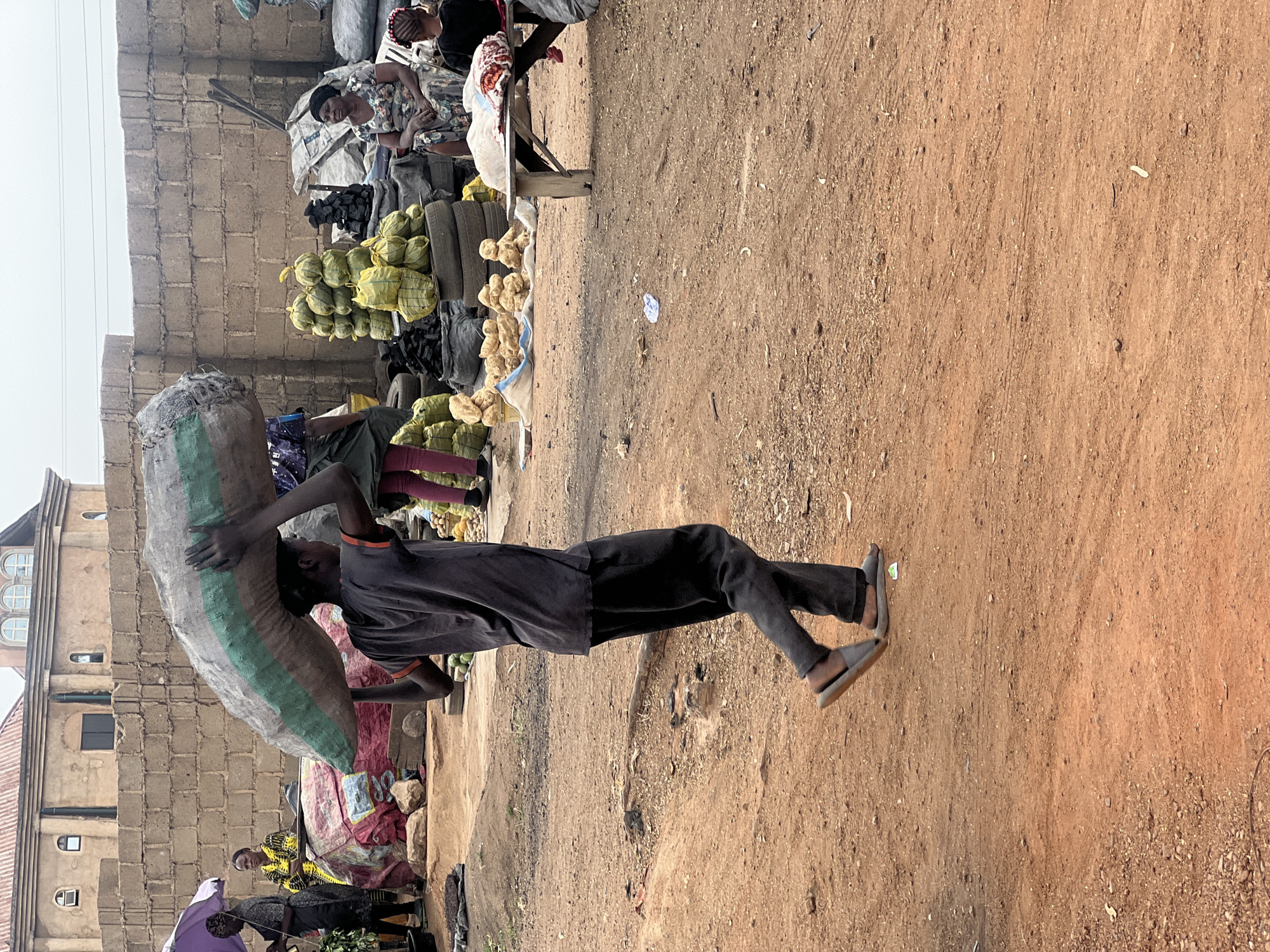 The image captures a vibrant market scene in Gura Top, Nigeria, on a bright, possibly warm day. The foreground features a young person, possibly male, walking from left to right across an uneven, reddish-brown dirt path. They are carrying a very large, heavy-looking sack, grey with a green stripe, balanced on their left shoulder and back, holding it with both hands. The person wears a dark t-shirt, dark trousers, and light-colored sandals.
Behind the moving individual, the scene unfolds into a bustling outdoor market. To the left, other market-goers or vendors are visible, including one person holding a purple umbrella. Further into the background, several market stalls are set up. One stall, towards the right, shows a woman in a patterned dress and headscarf, smiling, tending to her goods. These goods are varied and include piles of black items, likely charcoal, numerous bundles wrapped in yellow fabric, and small, light-colored root vegetables or produce arranged on the ground. Several dark, possibly old tires are also stacked near the stalls.
The market stalls are rudimentary, with wooden frames and fabric coverings, positioned against a backdrop of light-brown, unrendered brick buildings, some featuring arched windows. The sky above is clear but appears hazy, suggesting either dust in the air or a high sun. The ground throughout the market area is unpaved, dusty, and well-trodden. No discernible text is visible on any signs or objects within the frame. The image conveys a sense of daily commerce and activity in an African market setting.