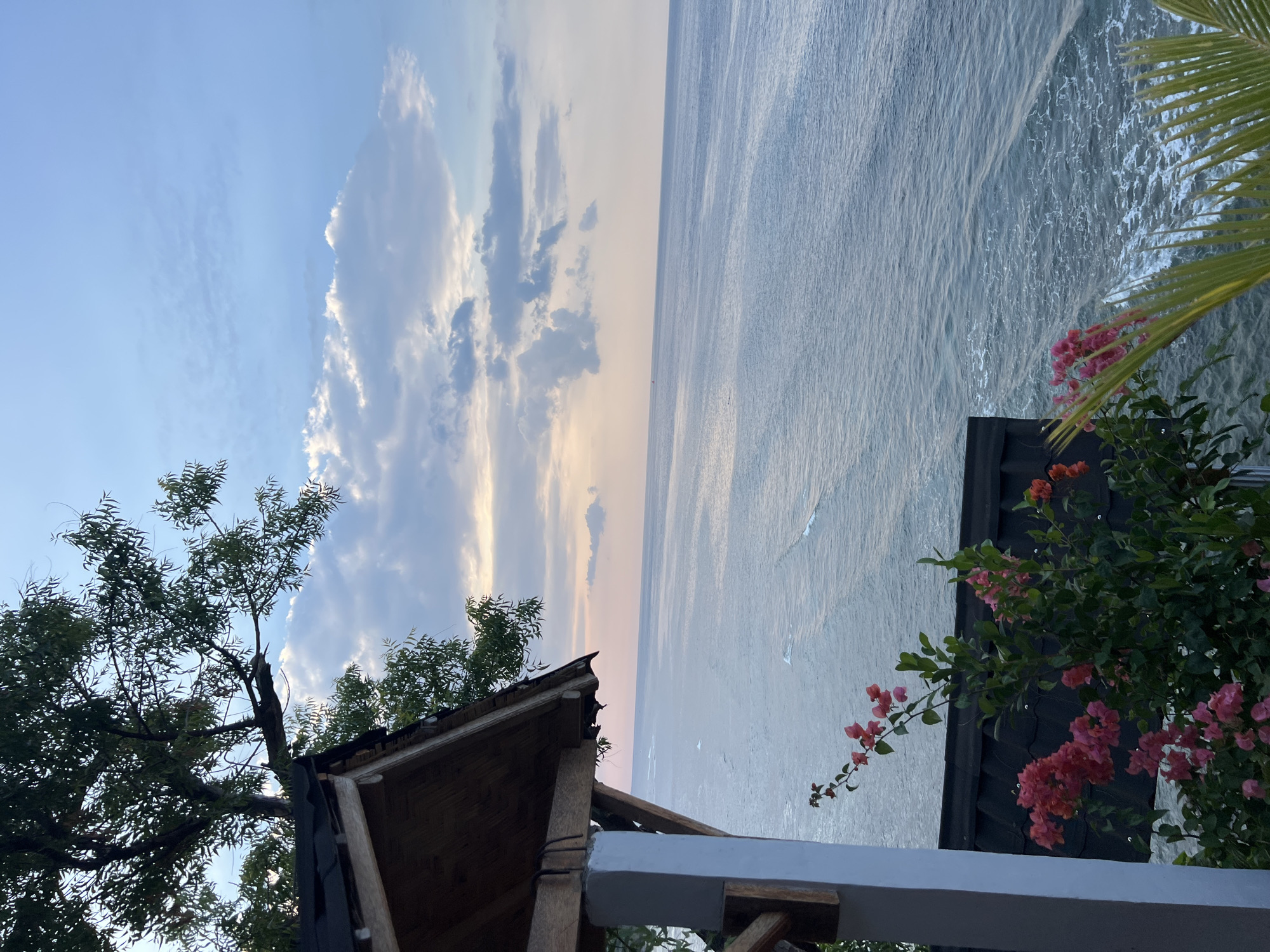 The image captures a serene view of the ocean from a vantage point in Abang, Indonesia. In the foreground, a small, wooden-roofed structure with a white pillar provides a partial framing of the scene. Lush green foliage and vibrant pink bougainvillea flowers cascade down from the structure, adding a touch of color to the composition. Beyond, the vast expanse of the ocean stretches to the horizon, its surface rippled by gentle waves that catch the soft light. The sky above is a canvas of pale blue, adorned with scattered clouds that are tinged with the warm hues of either sunrise or sunset, creating a tranquil and picturesque atmosphere. No people or animals are visible in the image.