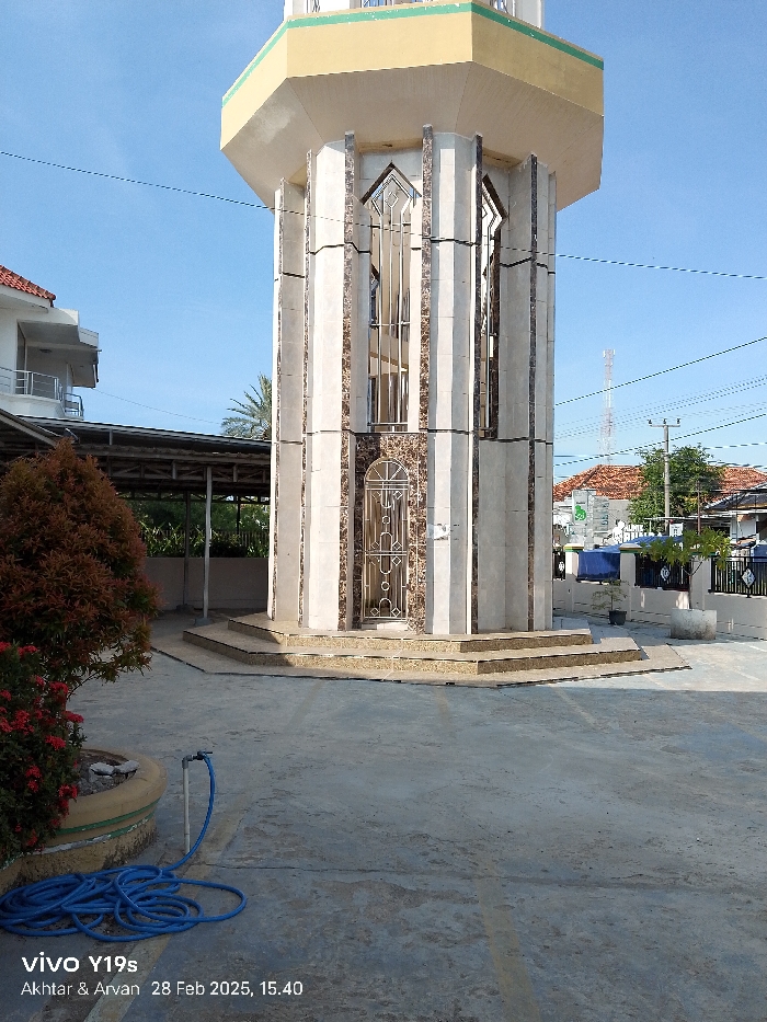 The image shows a tall, octagonal tower made of white stone and brown tile with a metal door on one side. The tower sits on a three-step platform made of the same stone and tile. The tower is located in the middle of a paved courtyard. There are two lines on the ground, one to the left and one to the right of the tower, both running from top to bottom. On the left side of the courtyard is a small bush with pink flowers in a yellow-brown planter. A blue hose lies coiled on the ground in front of the planter. The tower is located in Kecamatan Anjatan, Indonesia, with coordinates -6.35 latitude and 107.955 longitude. There is a blue tarpaulin covering a structure in the background, partially obscuring a building. The sky is a clear blue. The photo was taken on a sunny day, with no people visible.