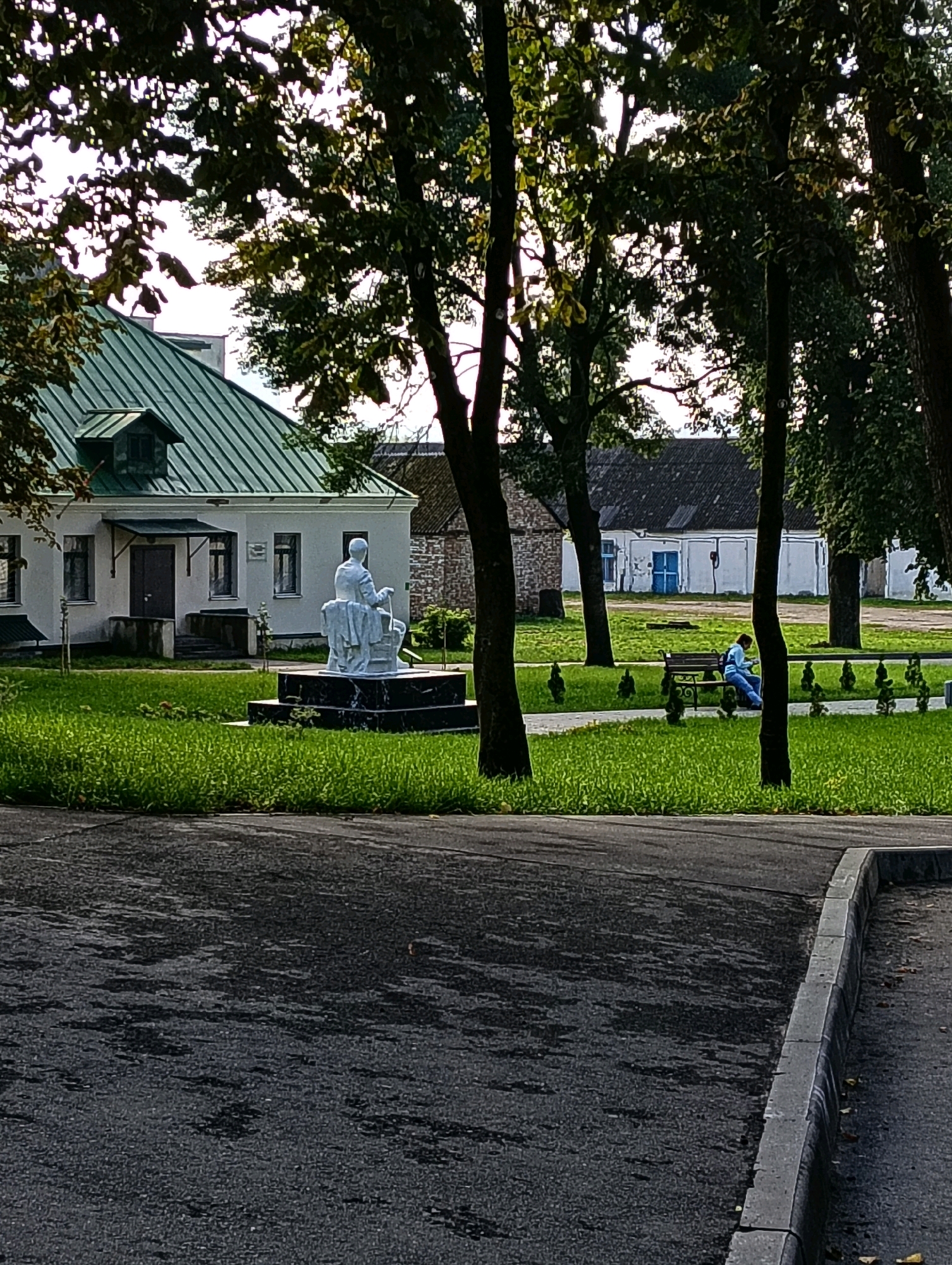 A person wearing a blue sweatshirt and blue jeans is sitting on a park bench to the right of the frame, looking down at a phone or book. They are in a park-like setting with green grass and several trees. To the left of the frame, there is a white building with a green roof and a dormer window. In front of the building is a white statue of a woman sitting and holding a bucket with a handle. The statue is on a black marble pedestal. In the background, there are more buildings with dark roofs and white walls. The ground in the foreground is asphalt. This image was taken in the city of Hrodna, Belarus.