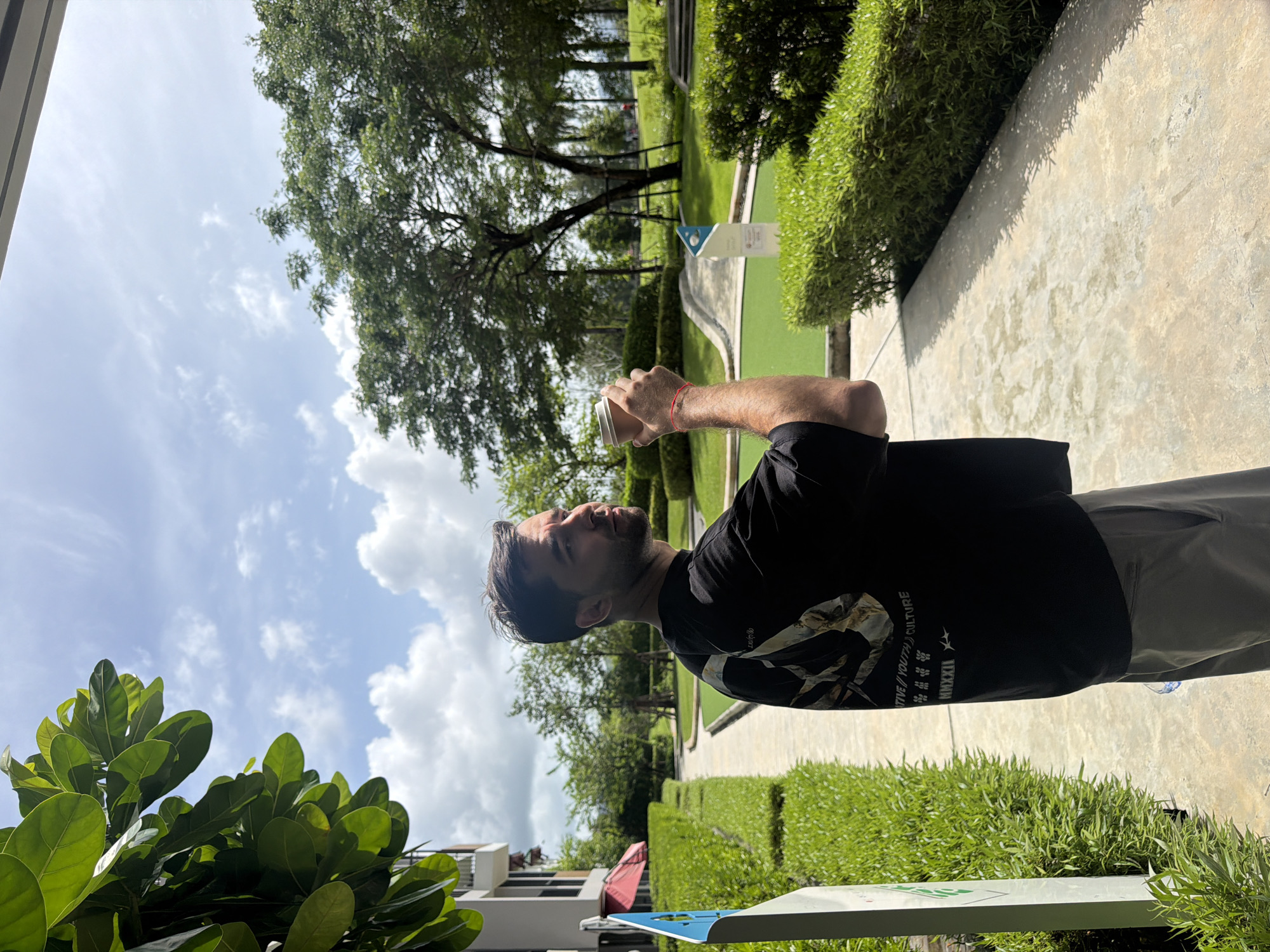 A Caucasian man with dark hair and a beard is holding a cup in his right hand. He is wearing a black t-shirt and gray pants. He appears to be in Choeng Thale, Thailand, on a paved path alongside a miniature golf course. The surroundings include trees, manicured hedges, a building, and a cloudy sky.