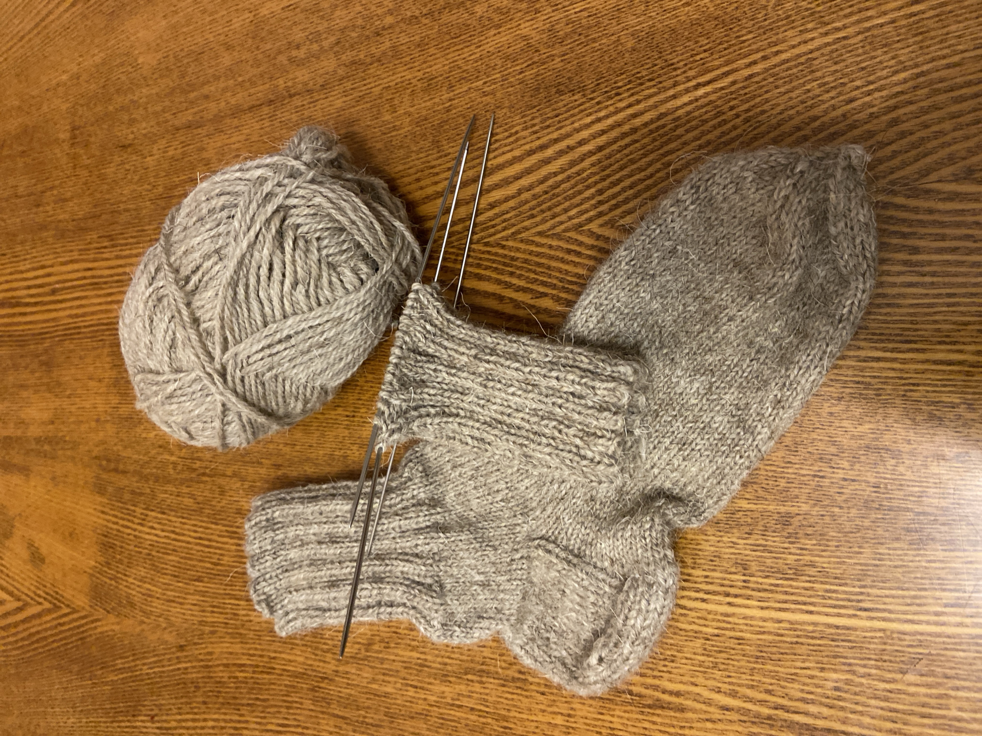 The image displays a pair of partially knitted gray socks, a ball of gray yarn, and knitting needles on a wooden surface. The knitting needles are positioned within the partially completed sock, indicating that the knitting process is in progress. The wooden surface appears to be a table with a visible wood grain pattern. The lighting suggests an indoor setting, possibly a home environment. There are no people present in the image. The scene is static and does not depict any specific action or interaction beyond the implied activity of knitting. The image is taken from a top-down perspective. The specific location of Aginskoye, Russia is not visually discernible from the provided image content.