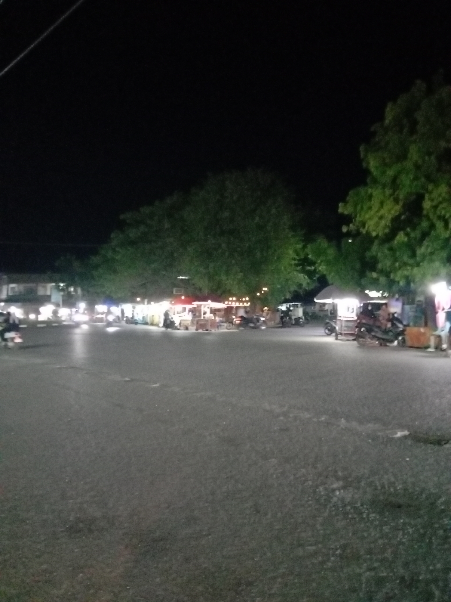 The image depicts a night scene in what appears to be a public square or open area in Keudekruenggeukueh, Indonesia. The ground is asphalt. There are scattered food stalls or carts, each illuminated. Motorbikes are parked or moving near the stalls. Trees are visible at the edge of the lighted area. Some buildings are visible in the background, illuminated by streetlights.