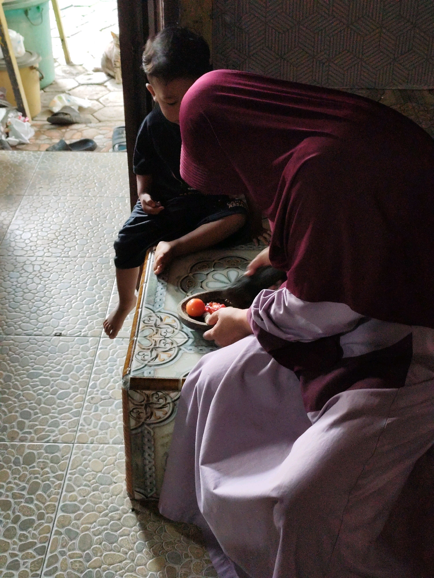 A young boy wearing a black t-shirt and dark pants sits on a table with his legs dangling. He is looking down. A person wearing a maroon hijab and a light purple dress, partially covering the boy's right side, is preparing food. On the table, a small bowl contains two tomatoes and some chopped ingredients. The table has a patterned decorative edge. The floor is tiled with a mosaic of small, rounded stones. In the background, a cat is visible in a dimly lit area with a green container and shoes on the floor. The scene is indoors, with natural light coming from an opening.