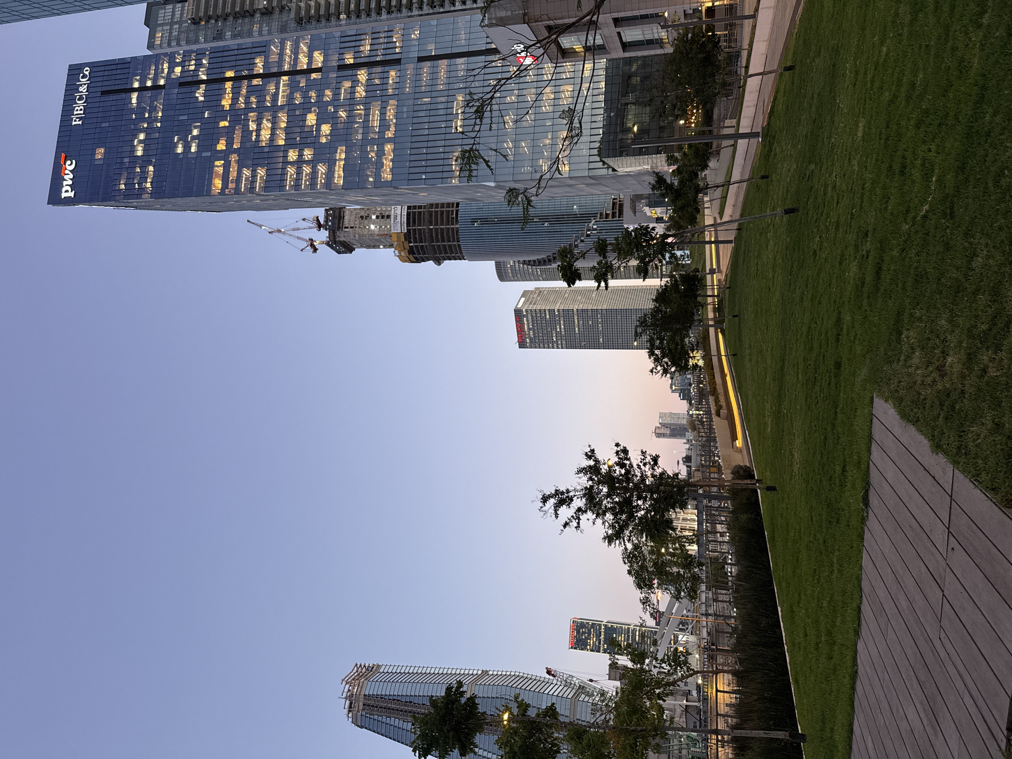The image depicts a cityscape in Tel Aviv, Israel at dusk. The sky is a gradient of light blue to soft pink near the horizon. Prominent in the foreground are modern skyscrapers, notably one with the PwC logo illuminated, indicating the presence of PwC Israel. A partially constructed building with cranes suggests ongoing development. A line of trees borders a green lawn adjacent to a wooden pathway. No people are visible. The scene suggests a modern, urban environment with active construction and landscaped public spaces.