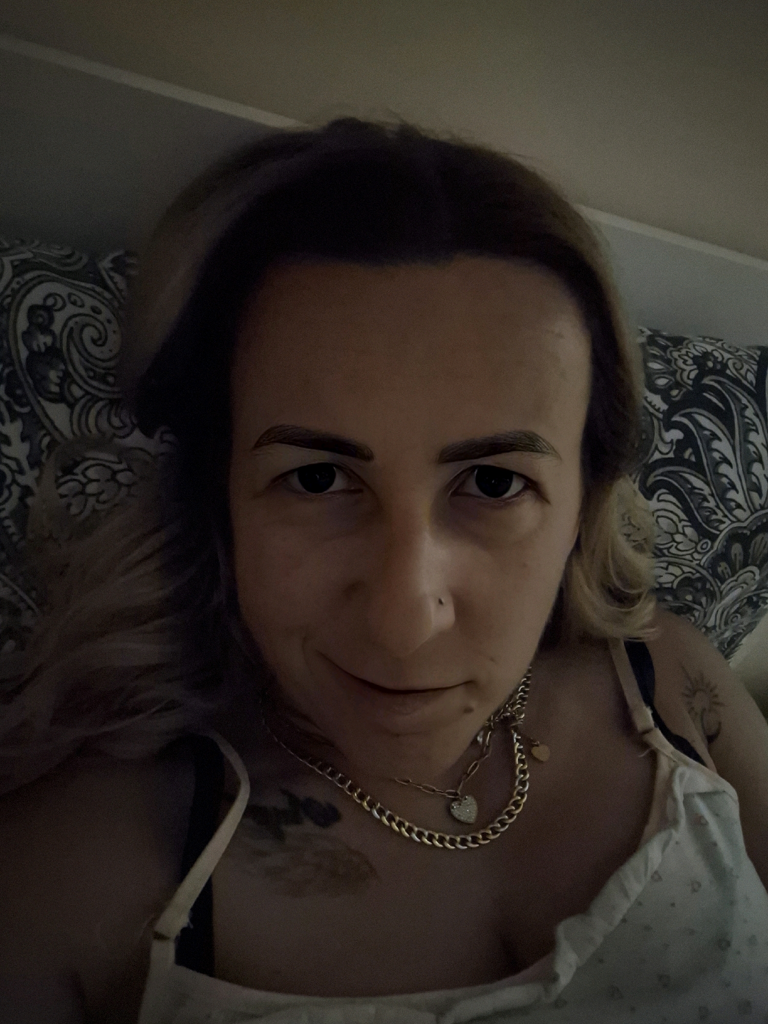 A close-up, waist-up, indoor selfie shows a person lying on their back, with their head resting on a pillow. The person has light brown hair, which is slightly messy and falls around their face. They have dark eyes, thin eyebrows, and a small nose piercing. The person is wearing a gold chain necklace with a heart-shaped pendant. They are also wearing a light-colored camisole or tank top, which has a delicate pattern on it, and appears to have a tattoo on their upper right shoulder, which is partially visible. The background consists of a pillow with a dark and white paisley pattern and a wall. The lighting is dim, suggesting it is nighttime or indoors with low light.