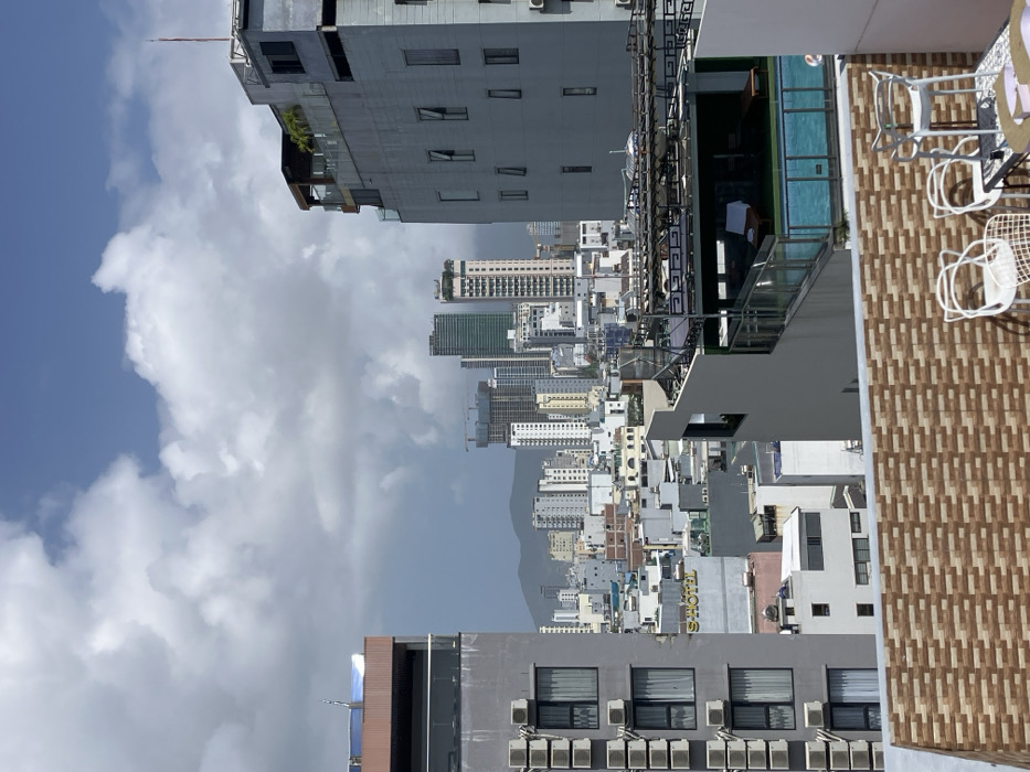 An aerial view captures an urban cityscape in Ngũ Hành Sơn Ward, Vietnam, from a high-rise rooftop perspective under a partly cloudy sky. In the foreground, a tiled rooftop terrace features a partial view of a small white table and three white metal chairs, two with distinctive arched backs. To the immediate left, a multi-story grey building presents a series of vertically stacked external air conditioning condenser units on its facade, with multiple windows visible. To the right, another multi-story grey building showcases a rooftop swimming pool with a glass safety barrier, beside which a dark lounge chair with a white towel is visible. Above the pool, an additional rooftop level is present with dark railings and sparse greenery.
Beyond these immediate structures, a dense urban landscape unfolds with numerous multi-story buildings of varying heights and architectural styles, predominantly white and grey. A yellow "S HOTEL" sign is visible on one of the mid-level buildings. In the distant background, several taller high-rise buildings dominate the skyline, including some under active construction with visible cranes and a distinctive green-colored high-rise. Faint mountain ranges are discernible on the distant horizon. The sky exhibits a mix of bright blue patches and large, white cumulus clouds, with darker cloud formations appearing towards the distant horizon. No individuals or specific actions are visible within the frame.