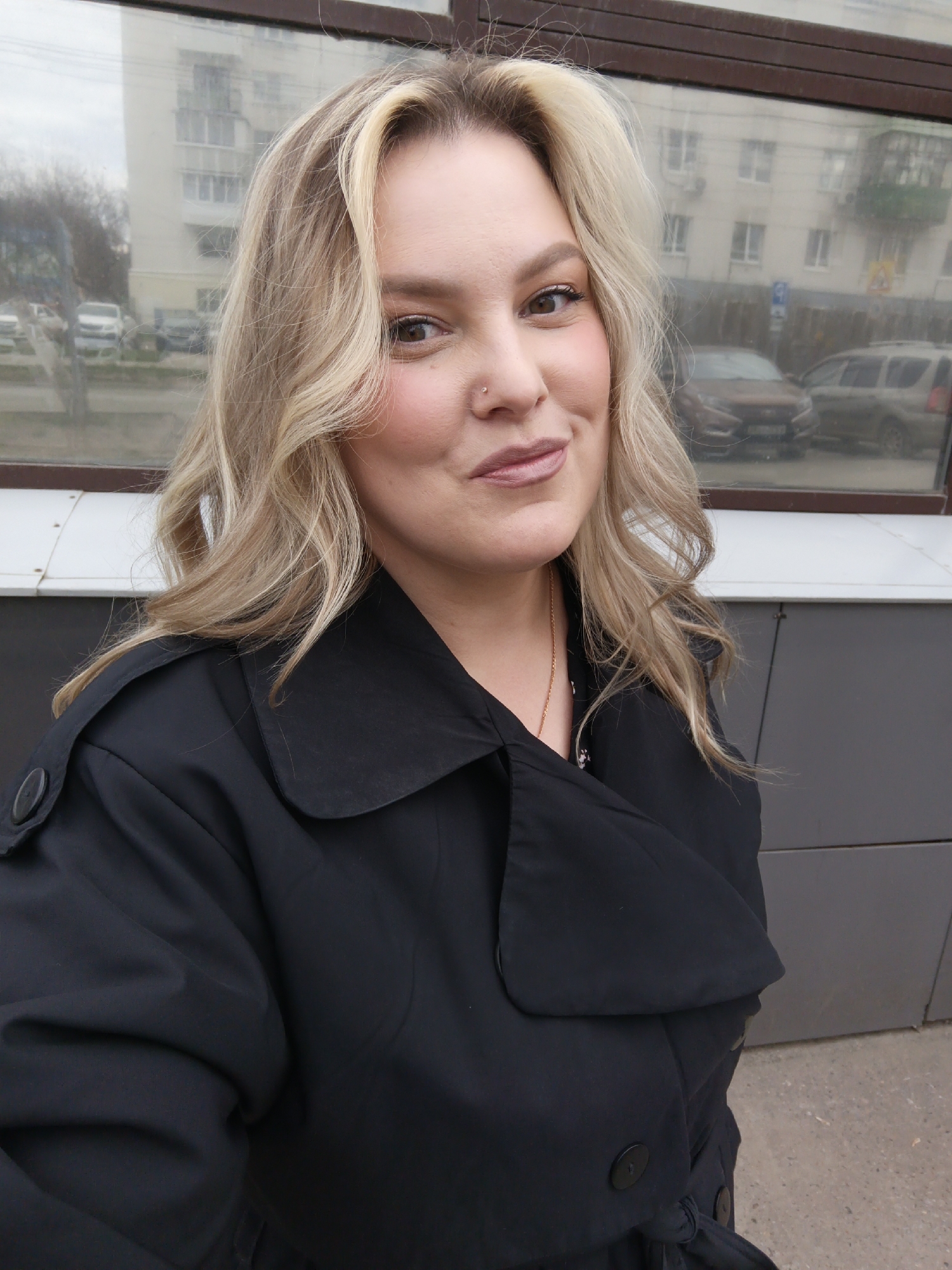 A woman is featured in a close-up, slightly high-angle portrait, likely a selfie. She has fair skin, light brown eyes, and a small silver piercing on her right nostril. Her wavy blonde hair, with darker roots and a prominent light streak near her face, frames her features. She wears natural-looking makeup, including blush, subtle eyeshadow, and light pink lipstick. Her expression is calm and pleasant, with a gentle, slightly playful smile as she looks directly at the camera.
She is wearing a dark, possibly black, trench coat or a similar collared jacket, visible from the chest down. The coat features wide lapels, large buttons, and shoulder epaulets. A thin gold chain is visible around her neck, beneath her coat.
The background is a reflection in a large glass surface, indicating she is outdoors in an urban setting. The reflection shows a multi-story residential building with numerous windows. Several parked cars are also visible, including a reddish-brown sedan and a grey SUV. Bare tree branches suggest a cooler time of year. Faint details of street signs, including what appears to be a blue parking sign, are also discernible. The lighting is soft and diffused, suggesting an overcast daytime. The scene captures a casual moment in Chaikovsky, Russia. No discernible text is visible in the image.