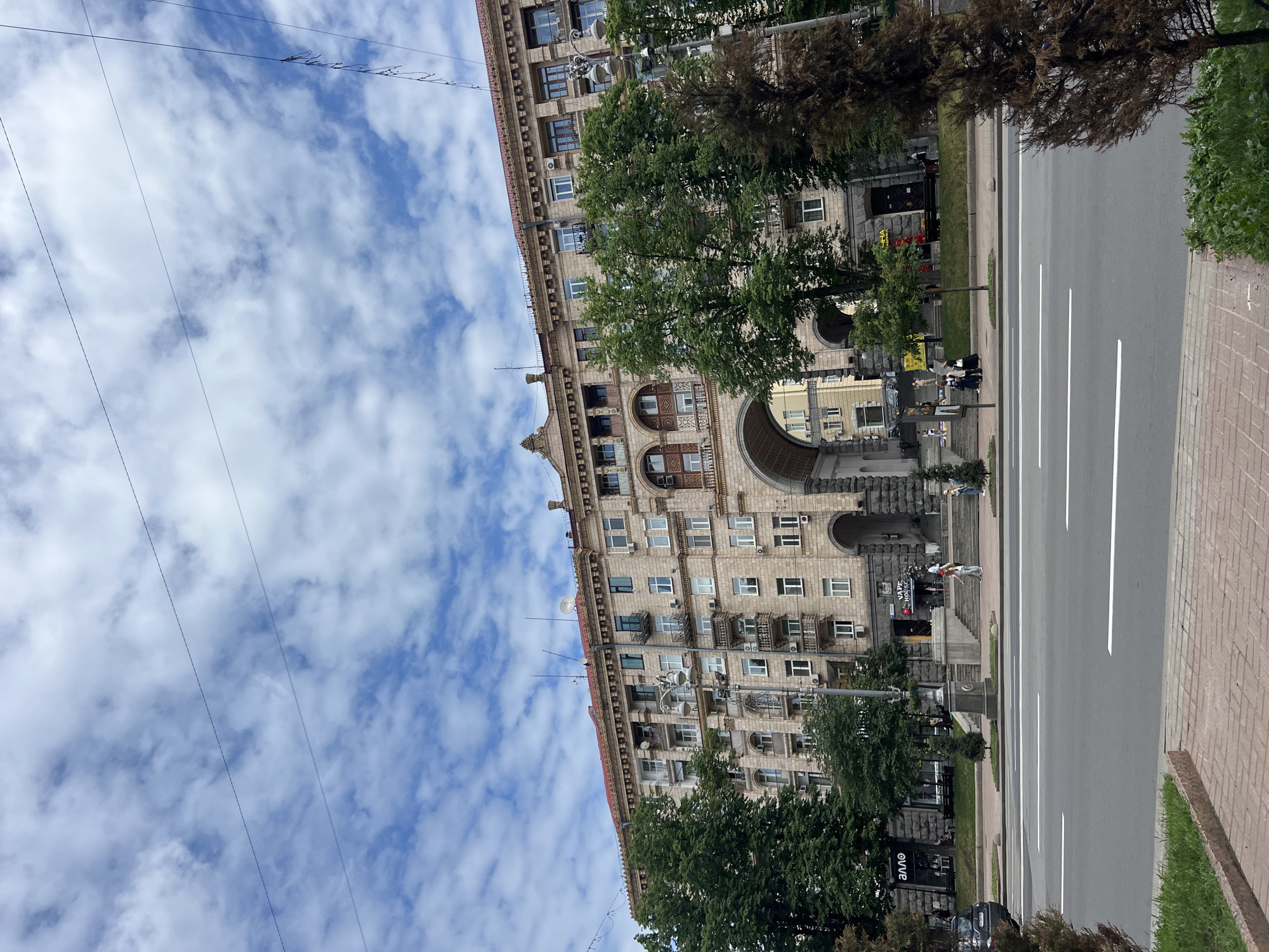 The image captures a street view in Kyiv, Ukraine, featuring a large, ornate building with a prominent archway. The building is multi-story, constructed of stone, with numerous windows and balconies. Trees line the sidewalk in front of the building, and a wide road with white lane markings runs through the foreground. The sky is partly cloudy. Several people are visible walking on the sidewalk and near the archway. A dark-colored car is parked on the left side of the street. Signs in Ukrainian are visible on some of the storefronts at the base of the building. The overall scene depicts an urban environment with a blend of architecture and greenery.