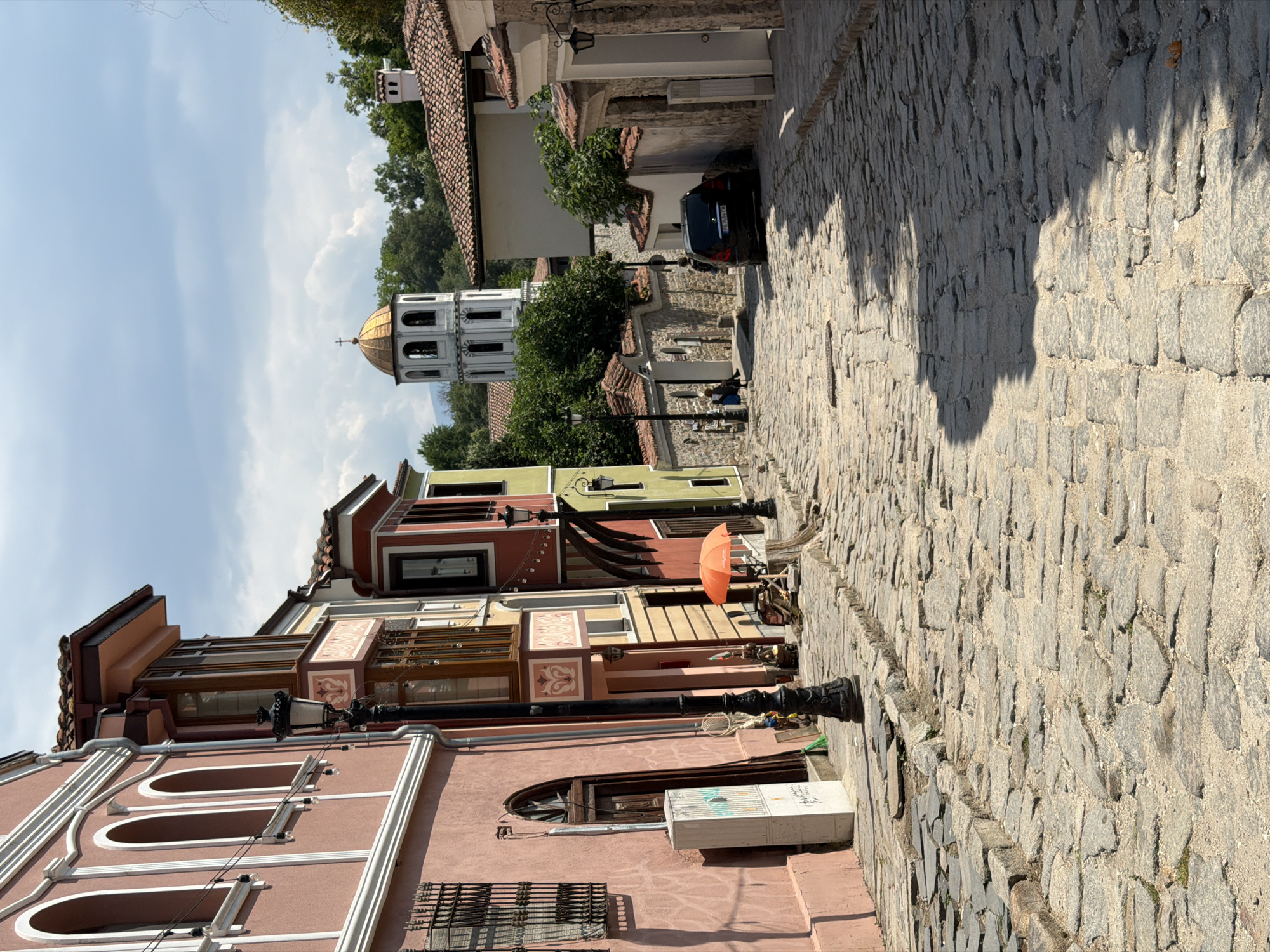 This image depicts a descending cobblestone street within the historic Old Town of Plovdiv, Bulgaria. The scene is illuminated by direct sunlight under a partly cloudy sky, casting pronounced shadows across the street.
On the left side of the street, a series of multi-story traditional Bulgarian Revival houses line the path. The prominent building in the foreground is painted a peach-pink hue, featuring three arched windows with white trim on its upper floor. Below, a window secured with a black metal grate and an arched wooden door are visible, alongside a white utility box adhered to the wall. A tall, ornate black street lamp post stands in front of this building. Further along the left, other buildings display varying colors including yellow, red, and light green, many with characteristic bay windows and tiled roofs. A small, open orange sun umbrella is positioned on the left side, near what appears to be a small vendor's setup.
The street itself is paved with irregular grey and beige cobblestones, indicating significant wear and age, and clearly slopes downwards towards the background. Patches of light and shadow create a mosaic pattern on its surface. On the right side of the street, low stone walls and the facades of more traditional houses with terracotta tiled roofs are visible. A black sedan with the Bulgarian license plate "PB 7654 CH" is parked on the right side of the street, facing away from the viewer. Two individuals are seated on a stone wall on the right side in the mid-ground, and two additional figures are observed walking further down the street.
In the distant background, prominently centered, stands the distinctive white bell tower of the Church of St. Constantine and Helena. The tower features multiple tiers of arched openings and is capped with a gleaming golden dome surmounted by a cross. Green trees and foliage are abundant behind the buildings and around the church tower, contributing to the historical and natural environment.