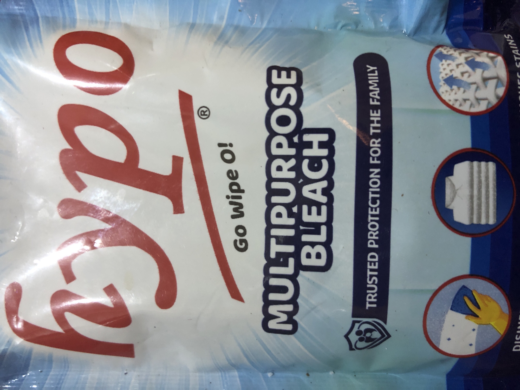 The image shows a close-up of a product bag, likely containing bleach. The brand name "hypo" is prominently displayed in large, red, stylized lettering across the top. Below this, the slogan "Go Wipe O!" is written in smaller black text. Further down, "MULTIPURPOSE BLEACH" is printed in large, bold, white letters with a dark blue outline. A dark blue band below this text contains the phrase "TRUSTED PROTECTION FOR THE FAMILY" in white lettering. To the left of this text, there is a shield-shaped logo containing a stylized representation of two adults and a child. At the bottom of the bag, there are three circular icons: one showing a gloved hand wiping a surface, another showing stacked white clothing, and a third depicting arrows rising from fabric, suggesting stain removal. The bag has a light blue background with abstract white and light blue patterns. The image is taken indoors, with some reflections and creases visible on the plastic packaging, indicating it is a product being photographed for commercial or informational purposes. There is no visible indication of a specific location within the city of Kabong, Nigeria, or any people present in the scene beyond the graphic representations on the product.