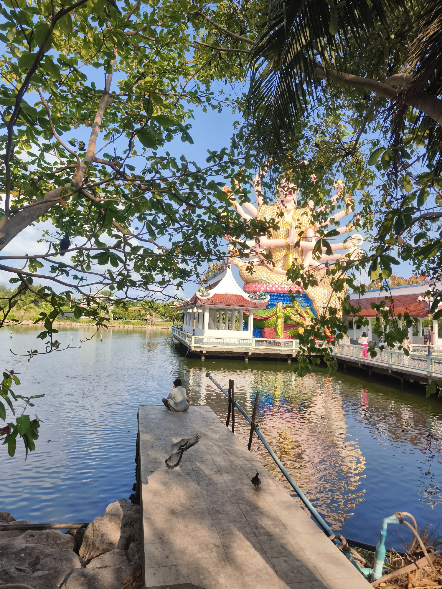 The image captures a scene in Baan Plai Laem, Thailand. A person sits at the end of a concrete pier on a lake. Two birds are visible on the pier, one mid-flight. In the background, a large, multi-armed statue sits atop a white and red building structure on an island in the lake. A walkway extends from the island towards the right side of the image. Trees frame the view, with branches and leaves visible in the foreground and top of the frame. The lake reflects the colors of the statue and buildings. The sky is blue with some clouds. Rocks line the edge of the pier where the person is sitting. A blue pipe is visible on the lower right edge of the pier.
