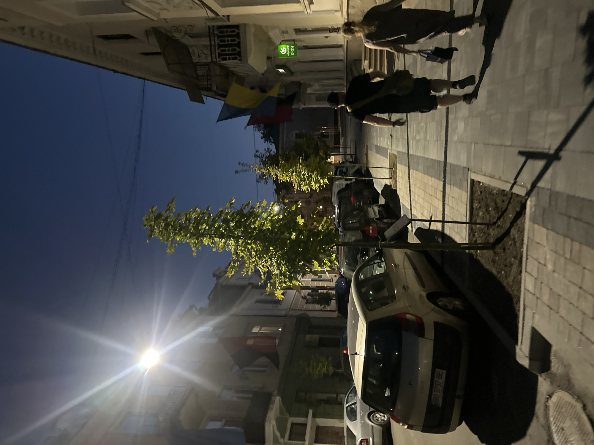 The image depicts a street scene in Ternopil, Ukraine, at dusk or early evening. The street is lined with buildings, one of which has a prominent bank sign that reads "efpbank 0-24". Ukrainian flags, red and yellow, are displayed on one of the buildings. A man and a woman are walking away from the camera on the sidewalk. The man is wearing a black t-shirt, black shorts, and a black cap, carrying a green bag over his shoulder. The woman is wearing a brown dress and carrying a dark bag. Several cars are parked along the street, including a silver Renault Scenic visible in the foreground with a license plate reading "BO 3737 BE". Streetlights are illuminated, casting long shadows. Trees are planted along the sidewalk. The sky is a deep blue, indicating the transition from day to night.