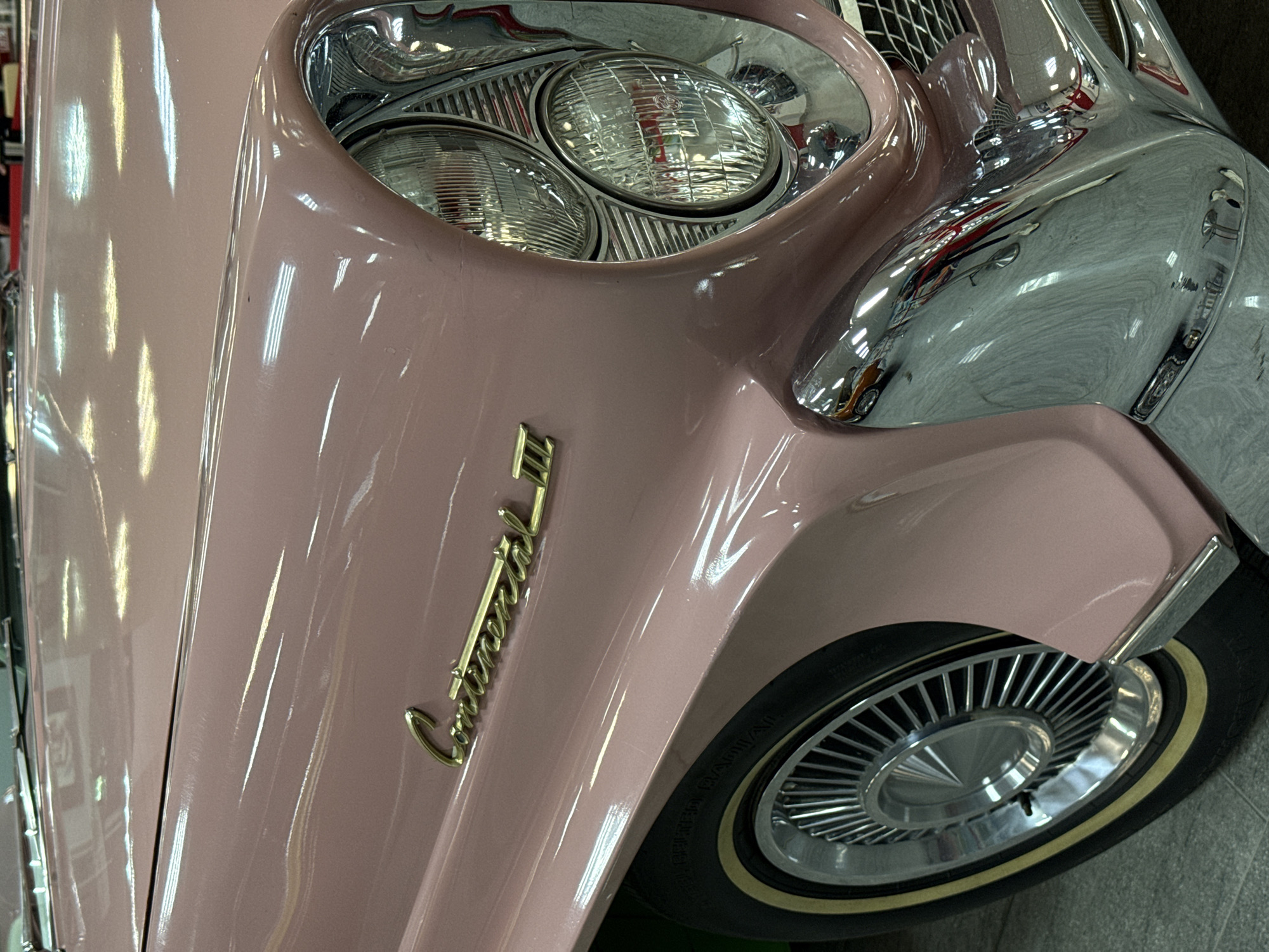 The image shows a close-up of the front of a pink classic car, specifically a Continental model. The prominent features include a stack of circular headlights surrounded by chrome housing, the golden "Continental" cursive emblem, the front wheel with chrome details and a white-wall tire. The car is likely indoors based on the reflections and artificial lighting. There is no information to determine if this image was taken in Sochi, Russia.