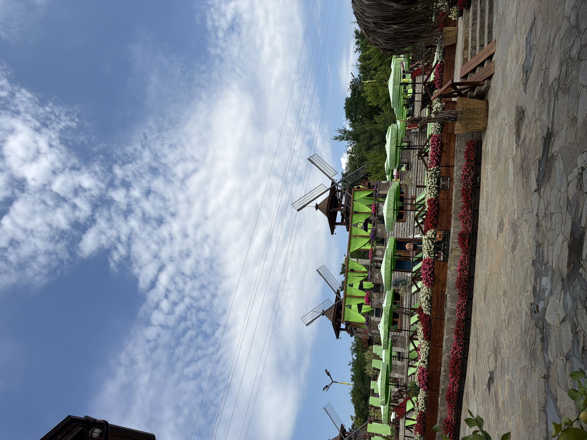 The image captures an outdoor seating area with a backdrop of several windmills, situated in the city of Gorna Malina, Bulgaria. The scene is set under a bright blue sky with scattered white clouds. In the foreground, a paved stone pathway leads into the seating area. Rows of wooden planters filled with vibrant red and pink flowers, along with white flowers, are strategically placed, creating a colorful border. Green umbrellas and lounge chairs are arranged on tiered platforms, suggesting a recreational or dining space. The windmills, a prominent feature, are constructed with wooden tops and large, angled blades, and are adorned with bright green fabric drapes. Several power lines are visible crisscrossing the sky in the upper portion of the image. Trees and lush greenery form a dense backdrop behind the structures. A person, appearing to be a male wearing dark shorts, is visible standing on a staircase on the right side of the image, interacting with the environment. To the far right, a thatched roof structure adds to the rustic ambiance. The overall impression is of a well-maintained outdoor venue designed for relaxation or leisure.