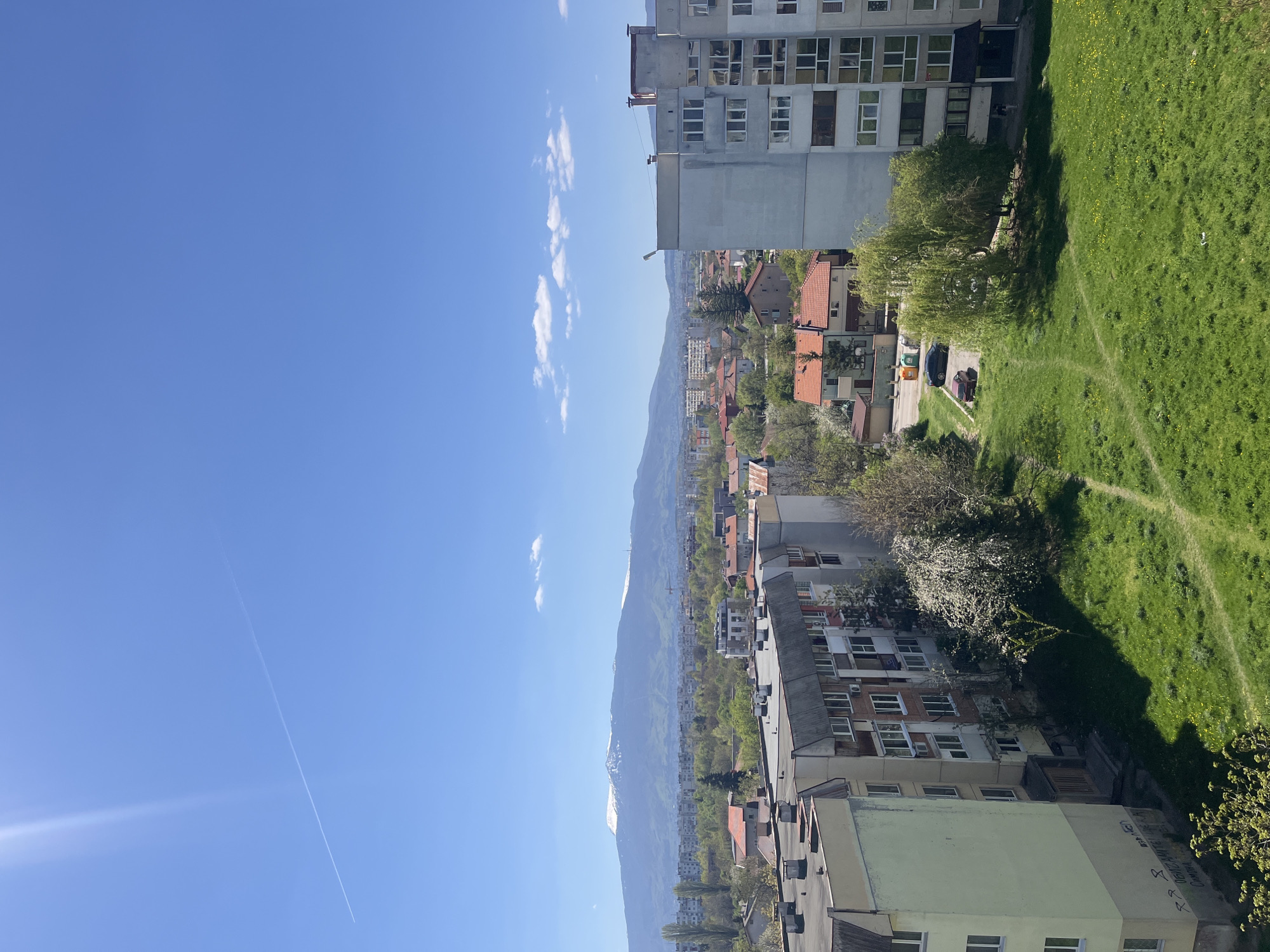 The image shows an elevated view of a city, likely Sofia, Bulgaria, on a clear day with a blue sky and some scattered clouds. A jet contrail is visible in the sky. The foreground includes buildings, some taller apartment blocks, and smaller residential houses with red tile roofs. A mountain range is visible in the middle distance. A green grassy area with visible paths is seen to the right. The light suggests it is daytime, possibly late morning or early afternoon. The scene appears peaceful and residential.