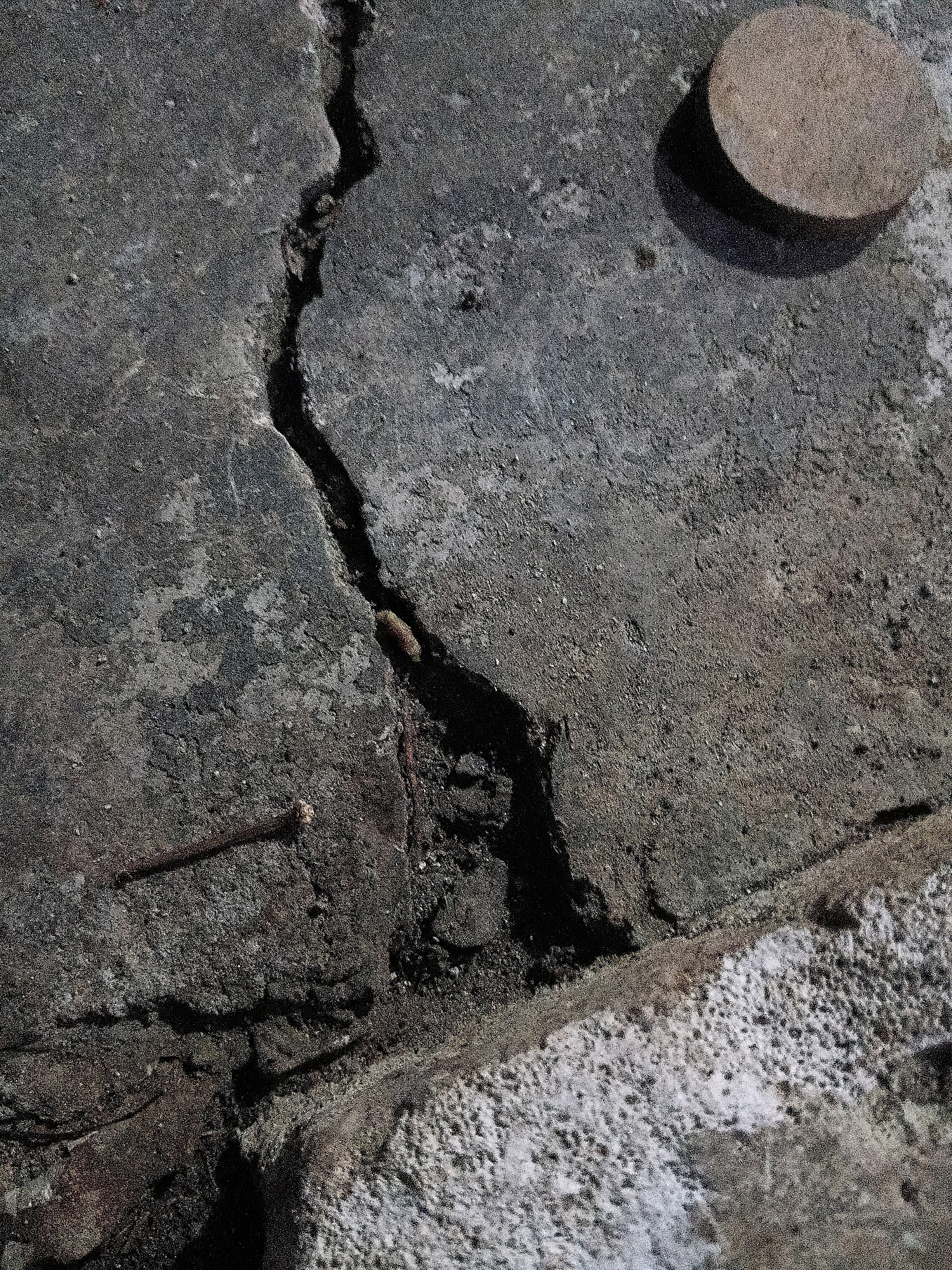 The image shows a close-up view of cracked concrete. A circular, light brown object, possibly a cap or a stopper, casts a shadow on the concrete in the upper right corner. A rusty nail is embedded in the concrete in the lower left quadrant, partially obscured by shadow. A dark, irregular crack runs diagonally through the center of the image, revealing dark soil and debris within its fissure. The texture of the concrete appears rough and weathered. The bottom edge of the image reveals a section of lighter, porous material adjacent to the concrete. The scene is devoid of any human presence or identifiable urban context beyond the material itself, making it impossible to determine its location within Cikal, Indonesia, or any other city.