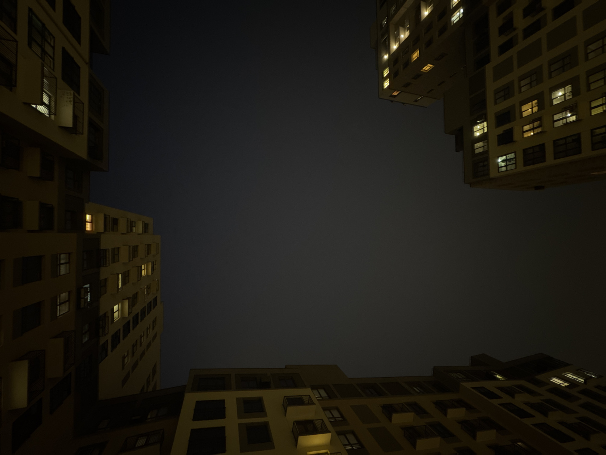 The image depicts a low-angle view of apartment buildings at night. The buildings are tall, with many windows, some of which are illuminated. The architecture appears modern and utilitarian. The sky is dark, suggesting it is nighttime. The buildings seem to form a perimeter around the viewer, enclosing the scene. The location is inferred to be Izhevsk, Russia, although this is not directly evident in the image itself. There are no people visible.