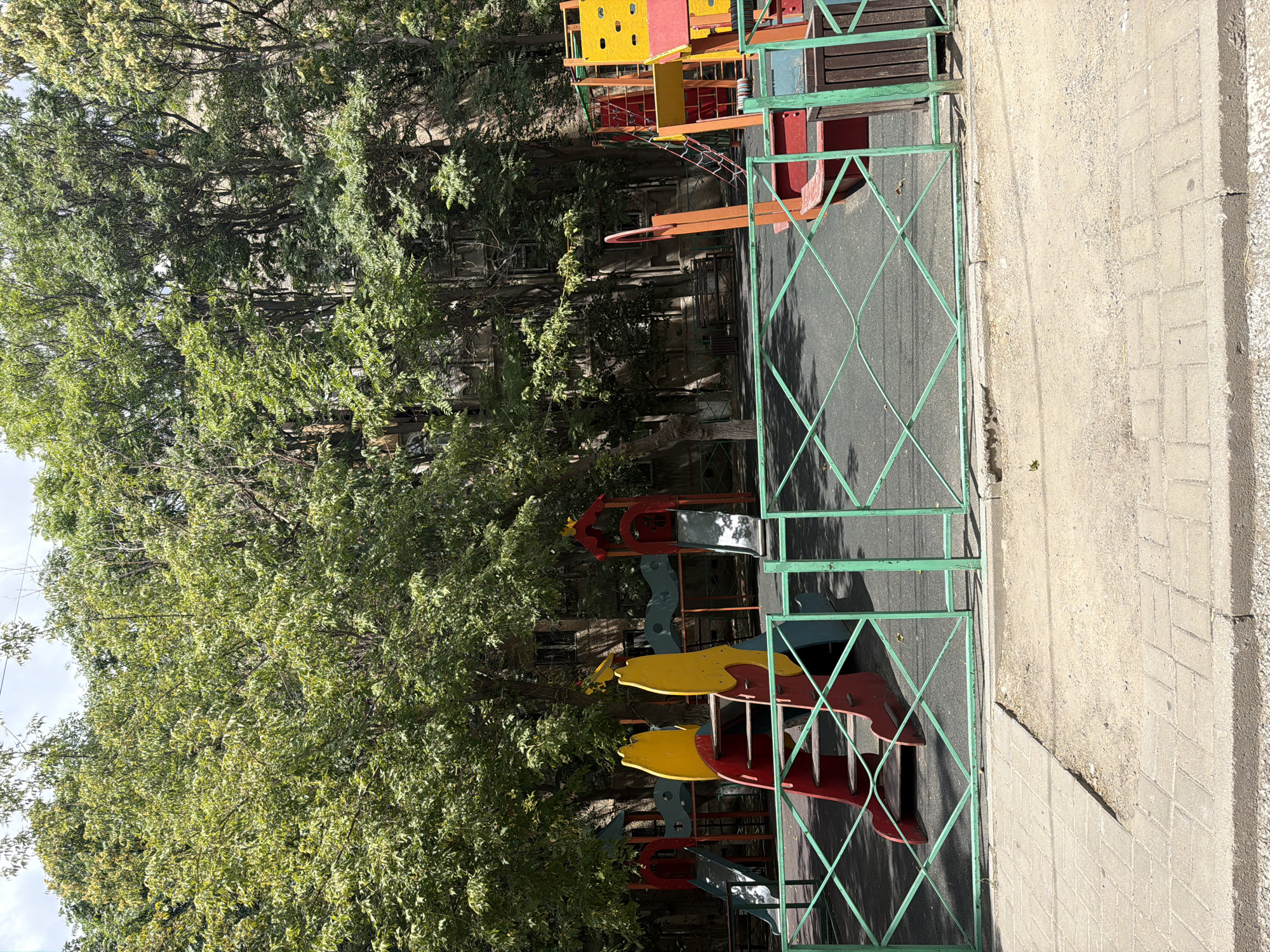 The image depicts a children's playground in Shymkent, Kazakhstan, framed by a green metal fence. The playground features several colorful slides and climbing structures, including a red and yellow slide with stairs, a blue and red structure with a slide, and a yellow and red climbing frame. The ground of the playground appears to be a dark, soft surface, possibly rubberized. Behind the playground, large, mature trees with dense green foliage create a backdrop, with the facades of apartment buildings visible through the leaves. A paved sidewalk is in the foreground, with a patch of dirt and gravel between the sidewalk and the playground fence. A wooden trash bin is situated to the right of the fence. The scene is illuminated by natural daylight, suggesting it was taken during the day. No people are visible in the image.