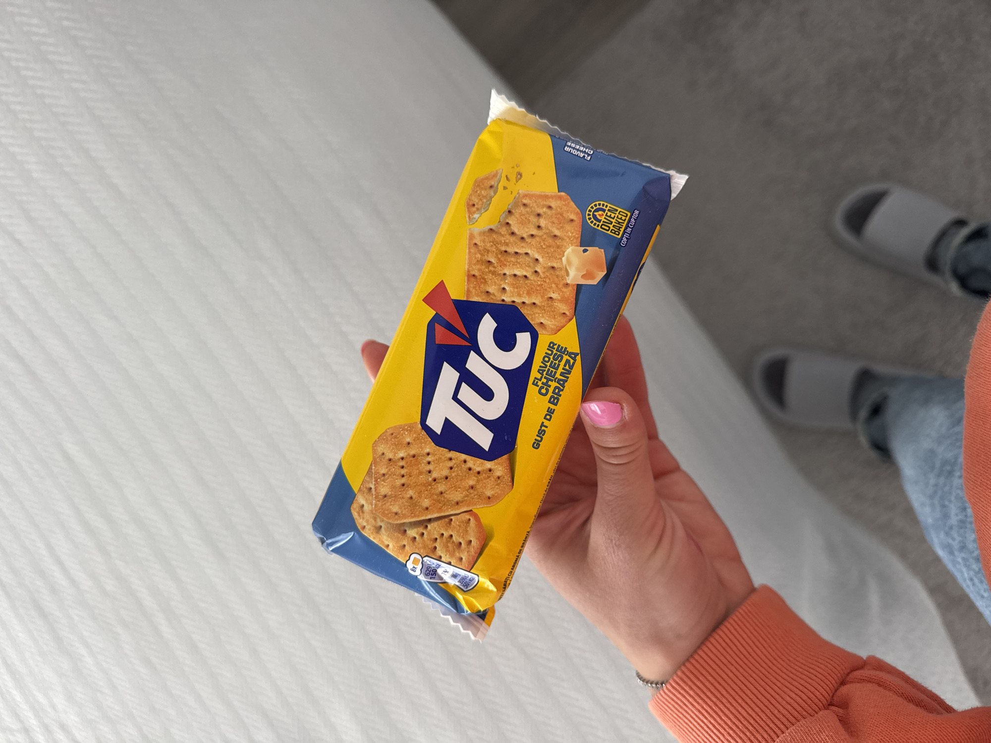 A hand holds a package of "TUC" brand cheese-flavored crackers, angled slightly towards the viewer. The package is predominantly yellow and blue, with images of the crackers. The hand has pink nail polish. In the background is a white textured surface and a person wearing slippers. The location is not specified, and there's no indication it's in Blagoevgrad, Bulgaria.