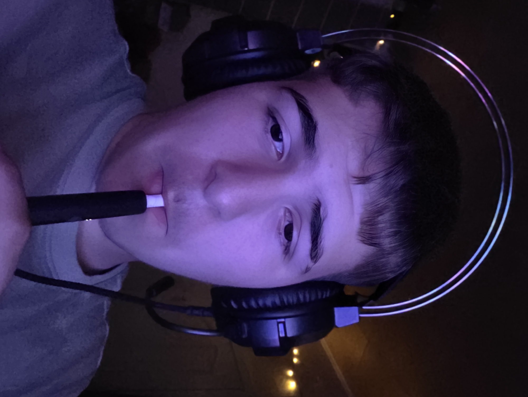 A young male, illuminated by purple light and wearing black headphones with a glowing band, is captured in a close-up selfie. He holds a dark cylindrical object with a white tip to his lips, suggesting he is smoking or vaping. The background is dimly lit, with some blurry yellow lights visible, hinting at an indoor environment, possibly a home or room. The image does not provide specific details about his location in Aleksandrovo, Bulgaria, or any other city.