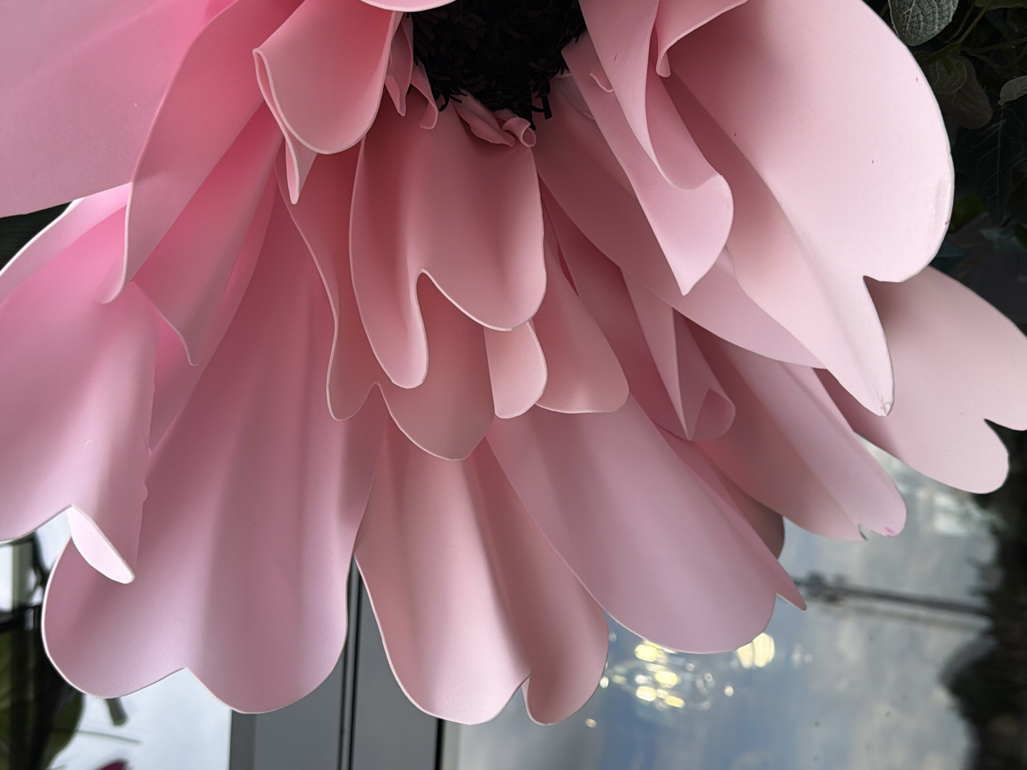 A close-up, low-angle shot shows the textured pink petals of a large, artificial flower. The petals have soft, rounded edges and are layered, creating a sense of depth. In the background, a window reflects a cloudy sky and some blurry lights, suggesting an indoor setting near a window. The flower's dark center is visible in the upper right portion of the frame. The scene is likely a decorative arrangement indoors, possibly in a shop or event space.