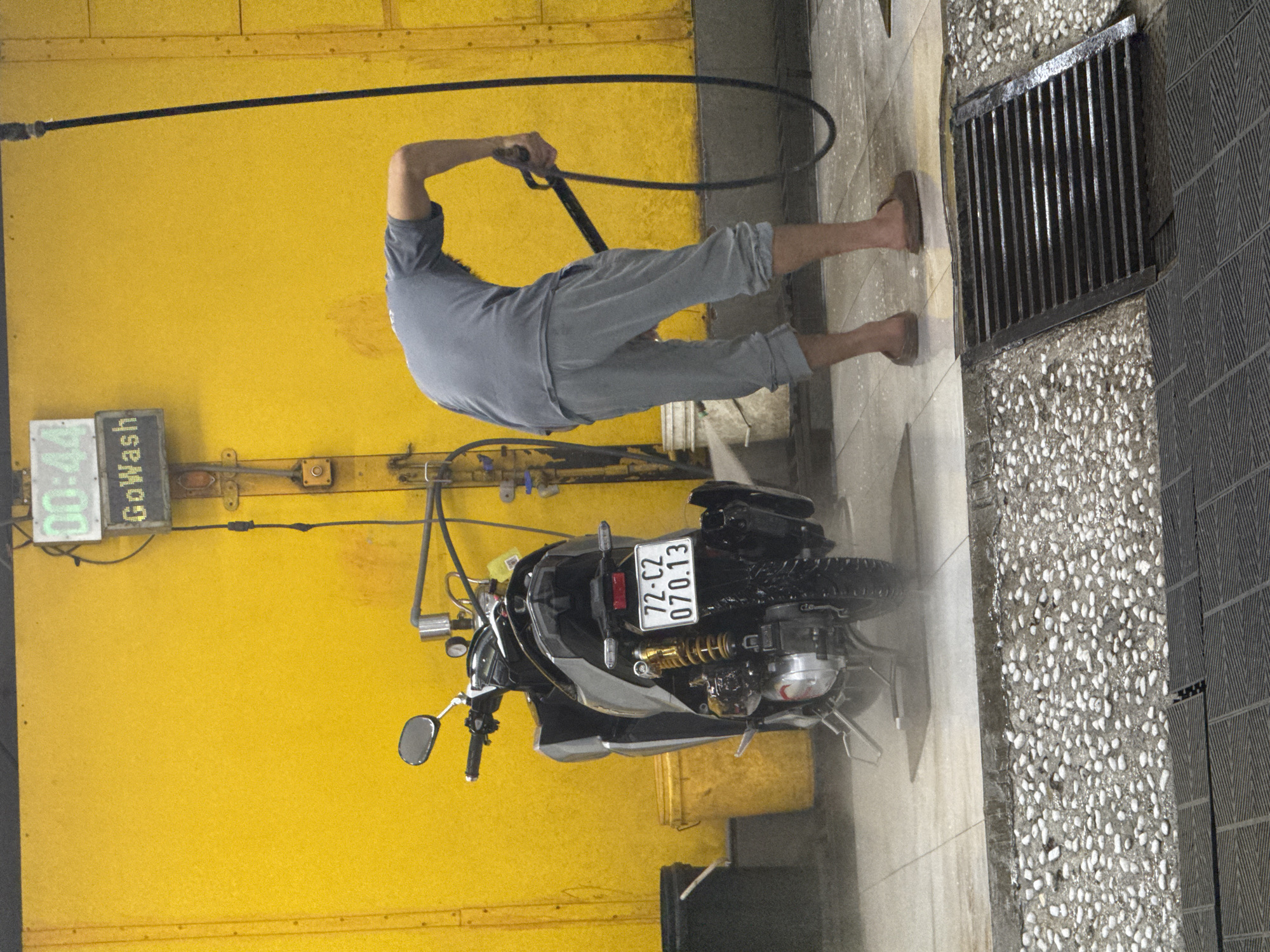 A person is washing a scooter with a high-pressure hose at a self-service car wash in Da Lat, Vietnam. The scooter is silver and black with a license plate reading "72-C2 070.13". The car wash has a bright yellow wall and a digital timer displaying "00:44". The ground is wet and there is a drain grate in the foreground. The person is wearing a grey t-shirt and grey pants, and appears to be barefoot. The environment is utilitarian, designed for cleaning vehicles.