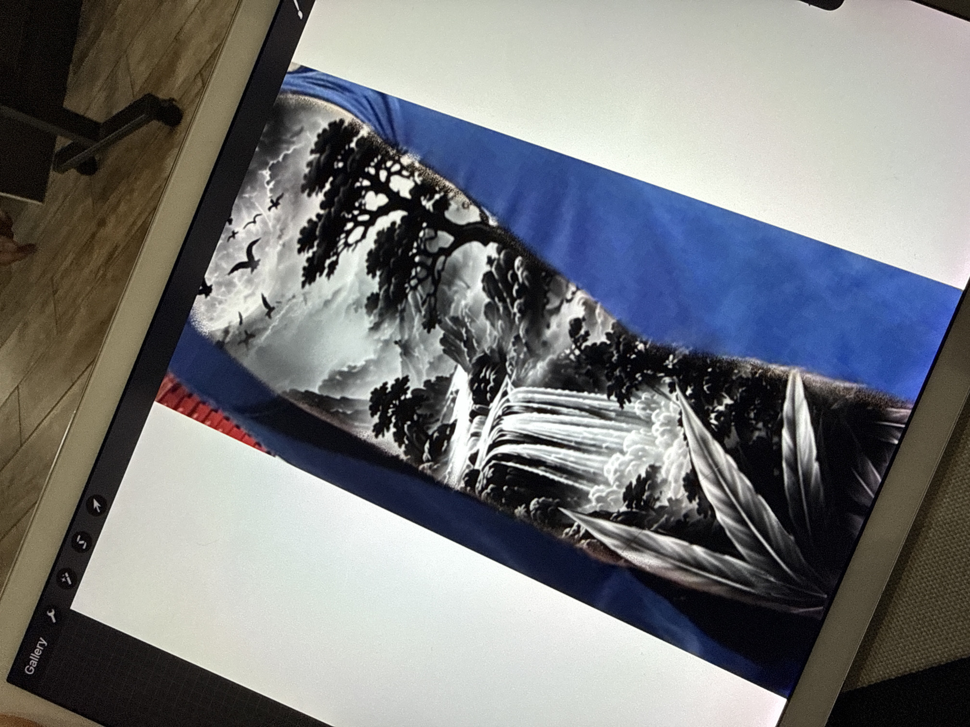 An indoor, close-up shot captures a person viewing a digital artwork on a light-colored tablet, likely in Sofia, Bulgaria. The tablet, held at an angle, displays a striking grayscale landscape design. This artwork, which strongly resembles a tattoo sleeve, features a dramatic scene with dark, gnarled trees, billowing clouds or mist, a powerful waterfall cascading down rocks, and several birds silhouetted against the lighter sky. Large, stylized leaves or feathers occupy the foreground on the right. The artwork is framed by solid blue areas, suggesting it might be a design mocked up on a limb or clothing.
The tablet's interface, visible on the left edge, includes a black sidebar with various tool icons (such as a brush, eraser, selection, and transform tools) and the word "Gallery" at the bottom, indicating a digital drawing application is in use. The background behind the tablet shows a warm-toned wooden wall with a visible door handle on the left, and a textured grey/brown fabric surface on the right, possibly from a couch or chair. The overall scene suggests a moment of artistic creation or appreciation in a comfortable interior setting.