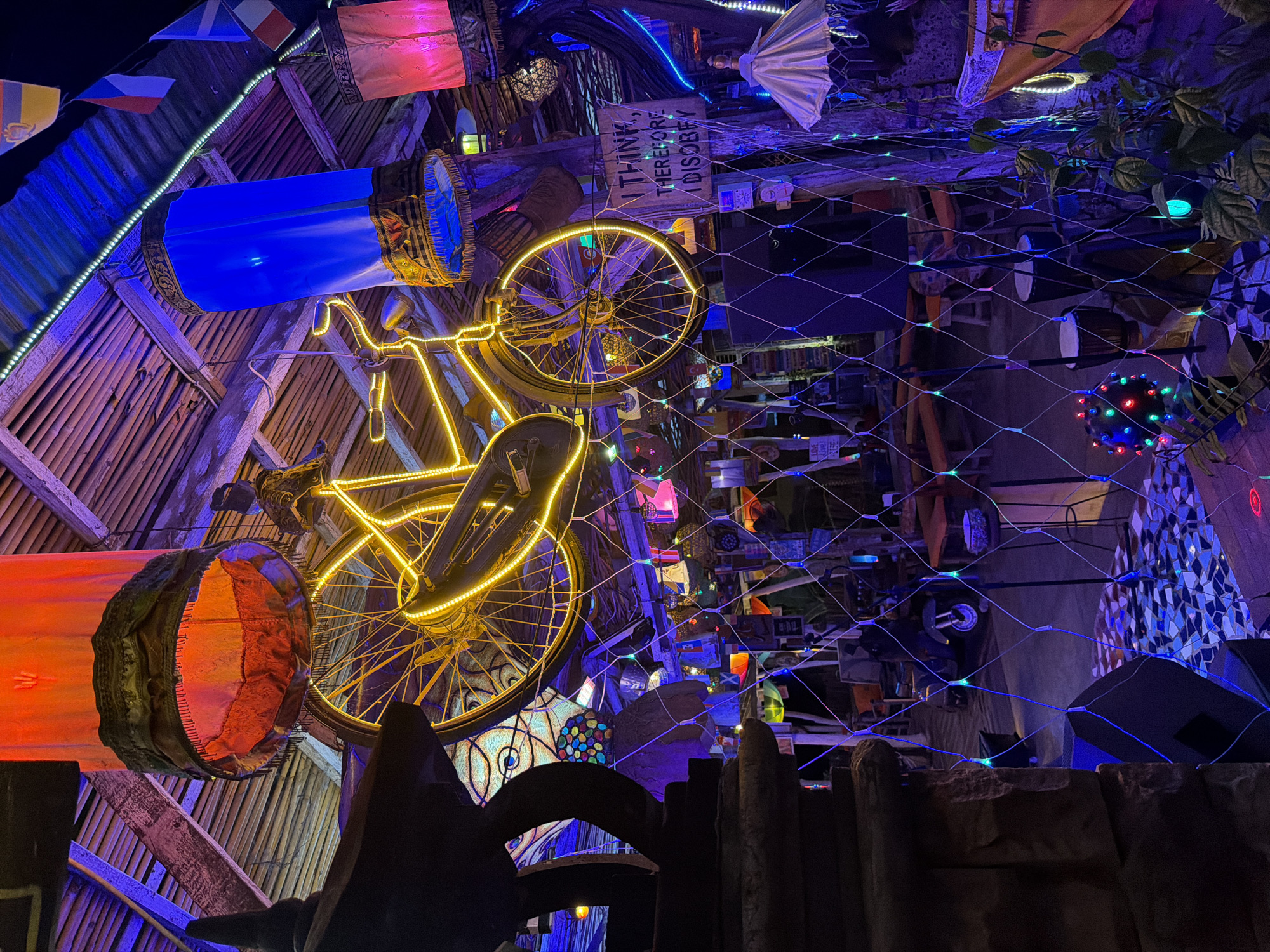 The image depicts the interior of a restaurant or bar in Banjar, Indonesia, characterized by a vibrant and eclectic decor. The ceiling is constructed from bamboo, with wooden beams supporting it, and adorned with various hanging lamps and decorations. Prominently featured is a vintage bicycle outlined in neon yellow lights, suspended from the ceiling. Other illuminated elements include a large blue cylindrical lantern, a red cylindrical lantern, and strings of multi-colored lights.
The space appears to be a seating area, with tables and chairs visible in the background, some with orange cushions. A net with embedded blue and green lights is strung across the foreground, creating a visual barrier. Among the decorative items are small flags hanging from the ceiling, a sign that reads "I THINK, THEREFORE I DISOBEY," a parasol-like object, and two bongo drums. Plants are visible in the lower right portion of the image. The overall ambiance is one of a dimly lit, bohemian, and festive establishment, likely designed to be an immersive and engaging experience for patrons. The lighting suggests it is nighttime or the interior lighting is quite low.