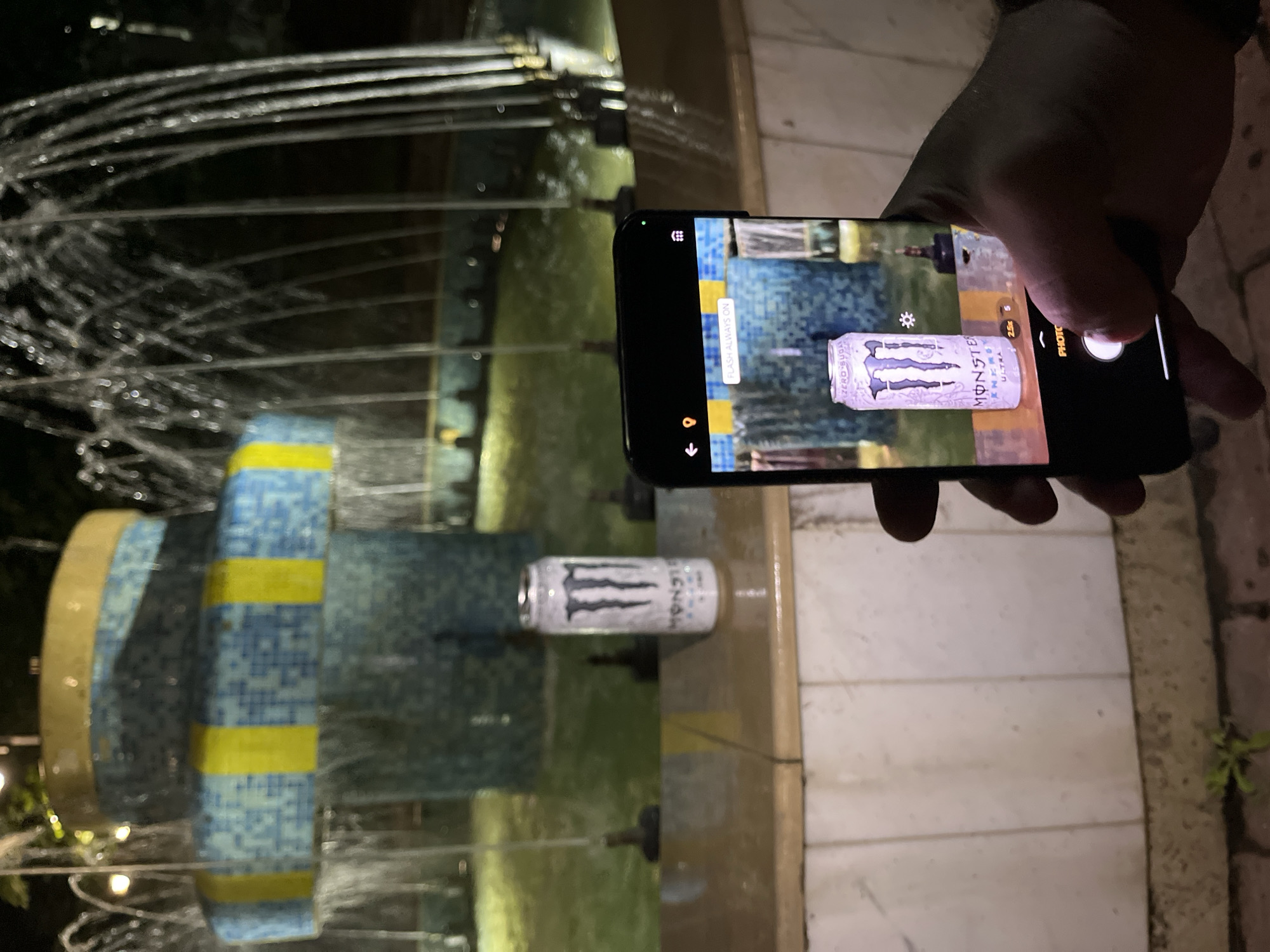 The image captures a nighttime scene in Veliki Preslav, Bulgaria, focusing on a person photographing a beverage can. In the foreground, a right hand holds a dark-colored smartphone, with its screen displaying the camera interface. The phone's camera is actively focused on a white Monster Energy Ultra Zero Sugar can, which stands upright on a beige tiled surface forming the edge of a water fountain. The phone screen shows "FLASH ALWAYS ON" and a "2.5x" zoom indicator.
The water fountain, located in the mid-ground, features multiple tiers adorned with blue mosaic tiles and yellow vertical stripe accents. Water actively cascades and sprays from several jets within the fountain, pooling in a basin where the water appears greenish due to ambient lighting. Several dark, small nozzles are visible within the water. The background consists of dark areas, with indistinct light sources. The ground beneath the smartphone consists of light-colored paving or tiling, with a small patch of green vegetation visible on the far left.