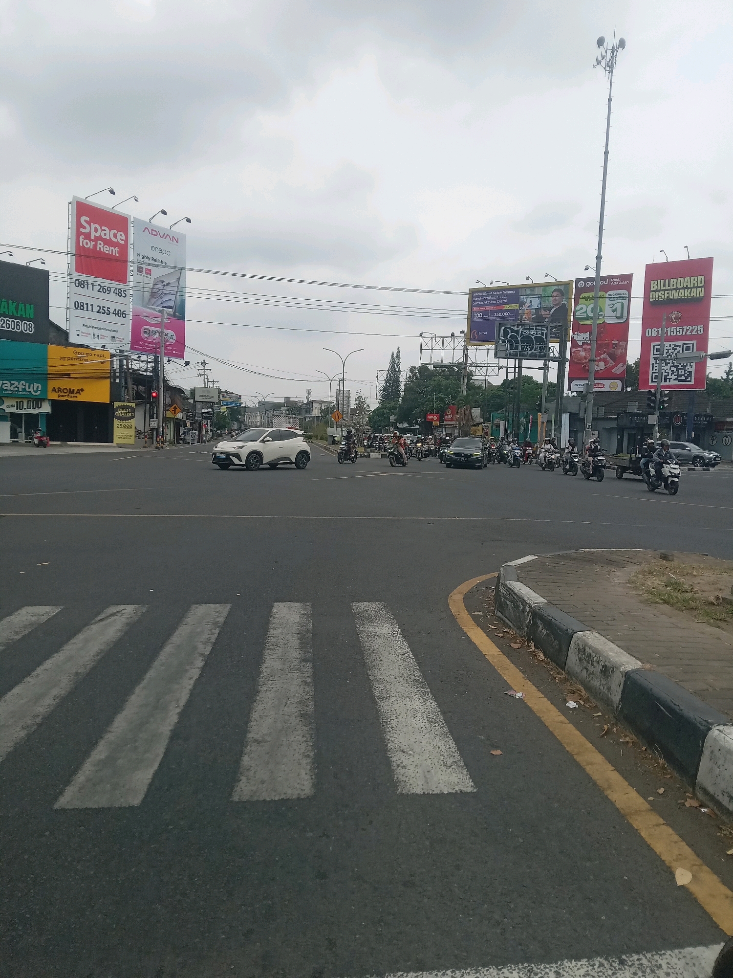 The image captures an outdoor urban scene, likely in Depok, Indonesia, characterized by a cloudy sky. In the foreground, a pedestrian crosswalk is visible on a paved road. The midground features a busy intersection with several vehicles, including a white SUV and numerous motorcycles. Traffic lights are present, indicating regulated movement. The background is populated by buildings, billboards, and a tall communication tower. On the left, a billboard reads "Space for Rent" with contact numbers. Another billboard advertises "ADVAN onepc Highly Reliable". Further to the right, a billboard states "BILLBOARD DISEWAKAN" (Billboard for Rent) with a phone number and a QR code. Various shops and businesses with signage can be observed along the roadside. The overall environment suggests a commercial and traffic-congested area.