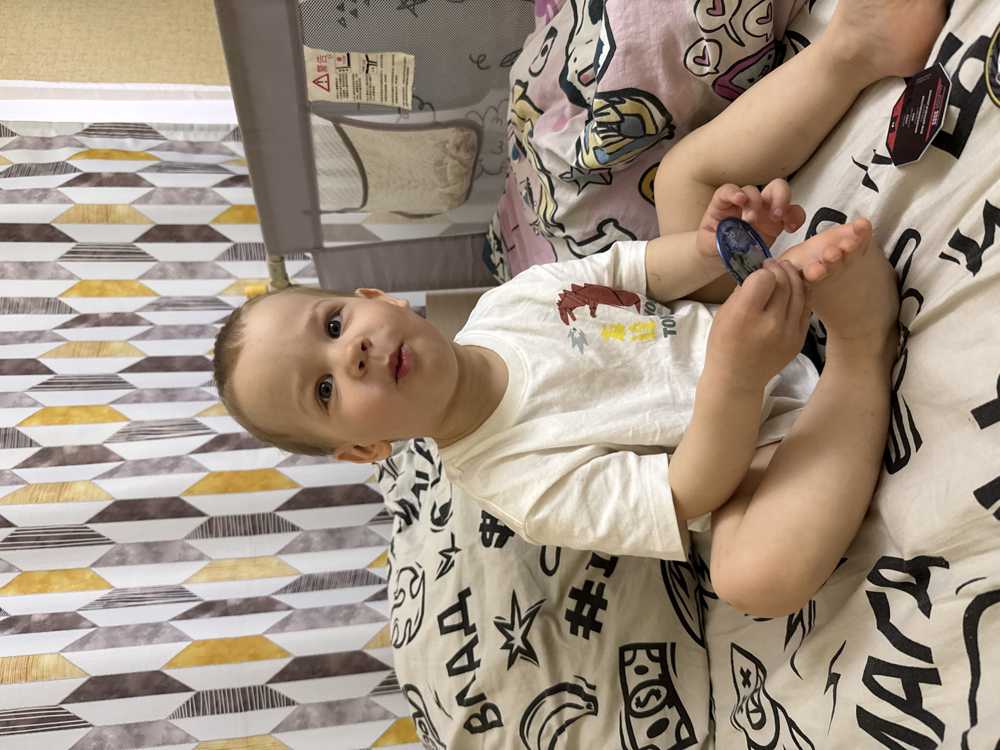A child is lying on a bed in an indoor environment. The child is fair-skinned with light brown hair and is wearing a white t-shirt. They are holding a small, round object in their hands and touching their foot with it. The bed has patterned bedding with black and white graphic designs. In the background, there is a playpen with mesh sides and patterned fabric. A portion of the floor is visible, which is covered with a patterned rug. The location is not clear from the image and can not be assumed to be Rusanovo, Russia.