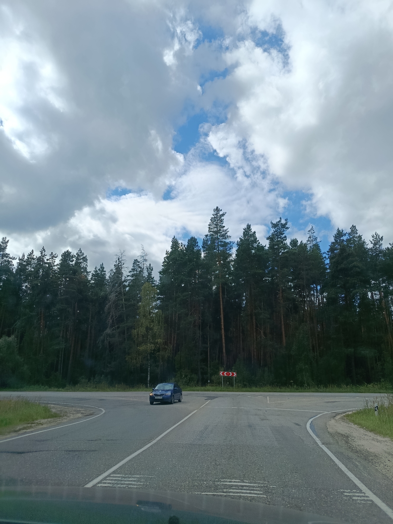 This is a photograph taken from inside a vehicle at a rural road intersection in Gribchikha, Russia. The scene is dominated by a dense forest of tall pine trees lining the background. The sky is partly cloudy with large, dramatic white clouds against a blue sky.
In the foreground, the asphalt road stretches towards the intersection. Lane markings are visible, including a solid white line on the right edge of the road and dashed white lines indicating lanes.
At the intersection, a dark blue passenger car is traveling on the road, appearing to be turning. A red and white directional sign with a chevron symbol is posted on the left side of the intersection, indicating a sharp turn.
The environment appears to be a natural, undeveloped area, with grass and vegetation growing along the sides of the road. The lighting suggests it is daytime, possibly late afternoon or early evening.