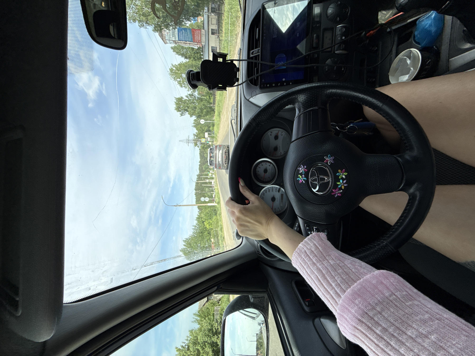 A first-person perspective from inside a car shows a person driving. The driver has their left hand on the steering wheel, and is wearing a light pink long-sleeved shirt. The steering wheel is black and features colorful flower decorations around the logo. Car dashboard components and a windshield-mounted phone holder are visible. Outside the windshield, the road ahead shows trees and buildings in the city of Chaikovsky, Russia.