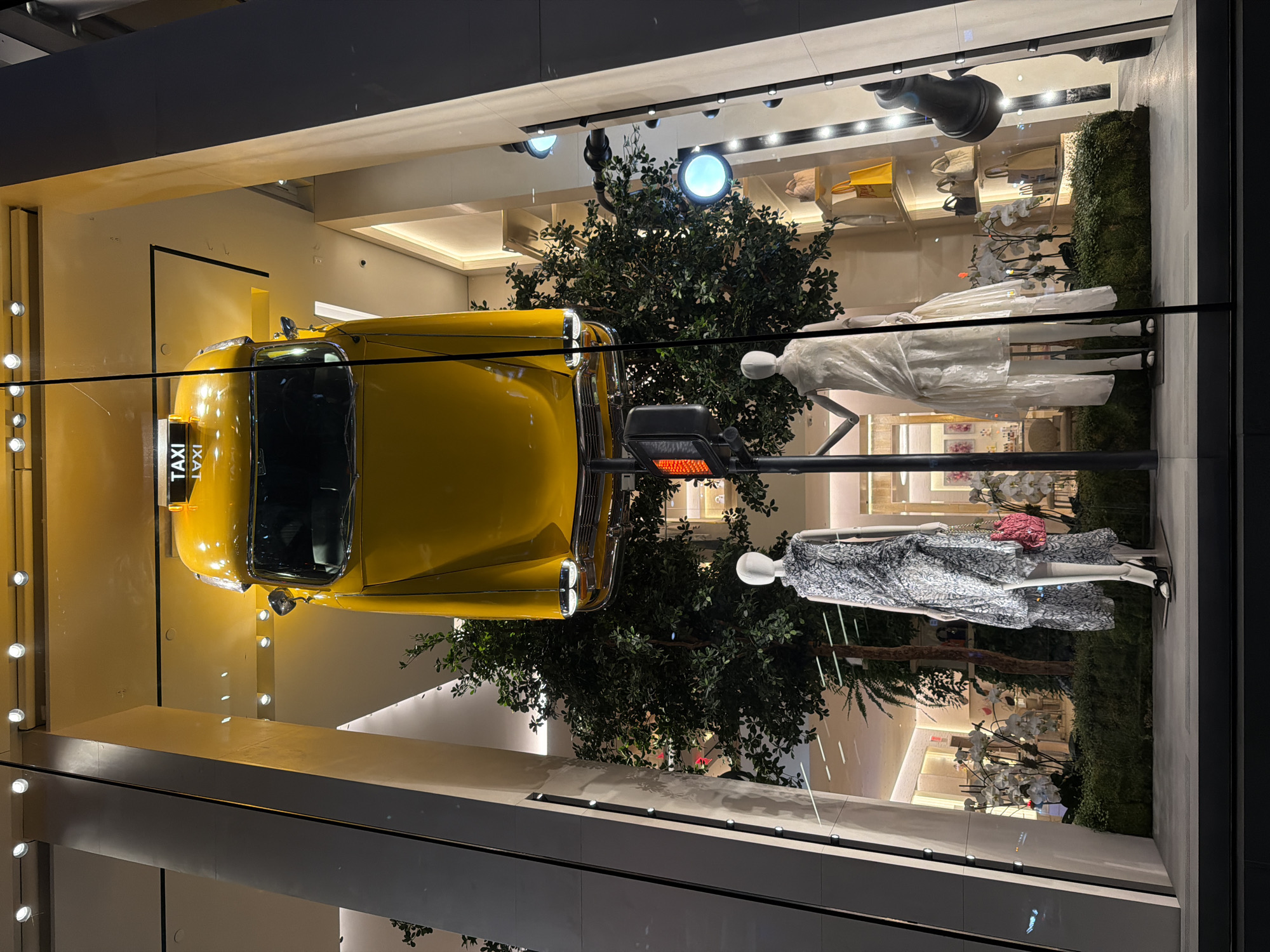 This image captures a New York City storefront window display featuring a yellow taxi suspended from the ceiling. The taxi's "TAXI" sign is visible. Below the taxi, two mannequins are displayed. The mannequin on the left wears a grey floral-patterned dress with a pink handbag. The mannequin on the right is dressed in a white, flowing gown. In the background, artificial trees and floral arrangements create a lush setting. Shelves with handbags and shopping bags are also visible behind the mannequins. The scene is illuminated by interior lighting and spotlights.