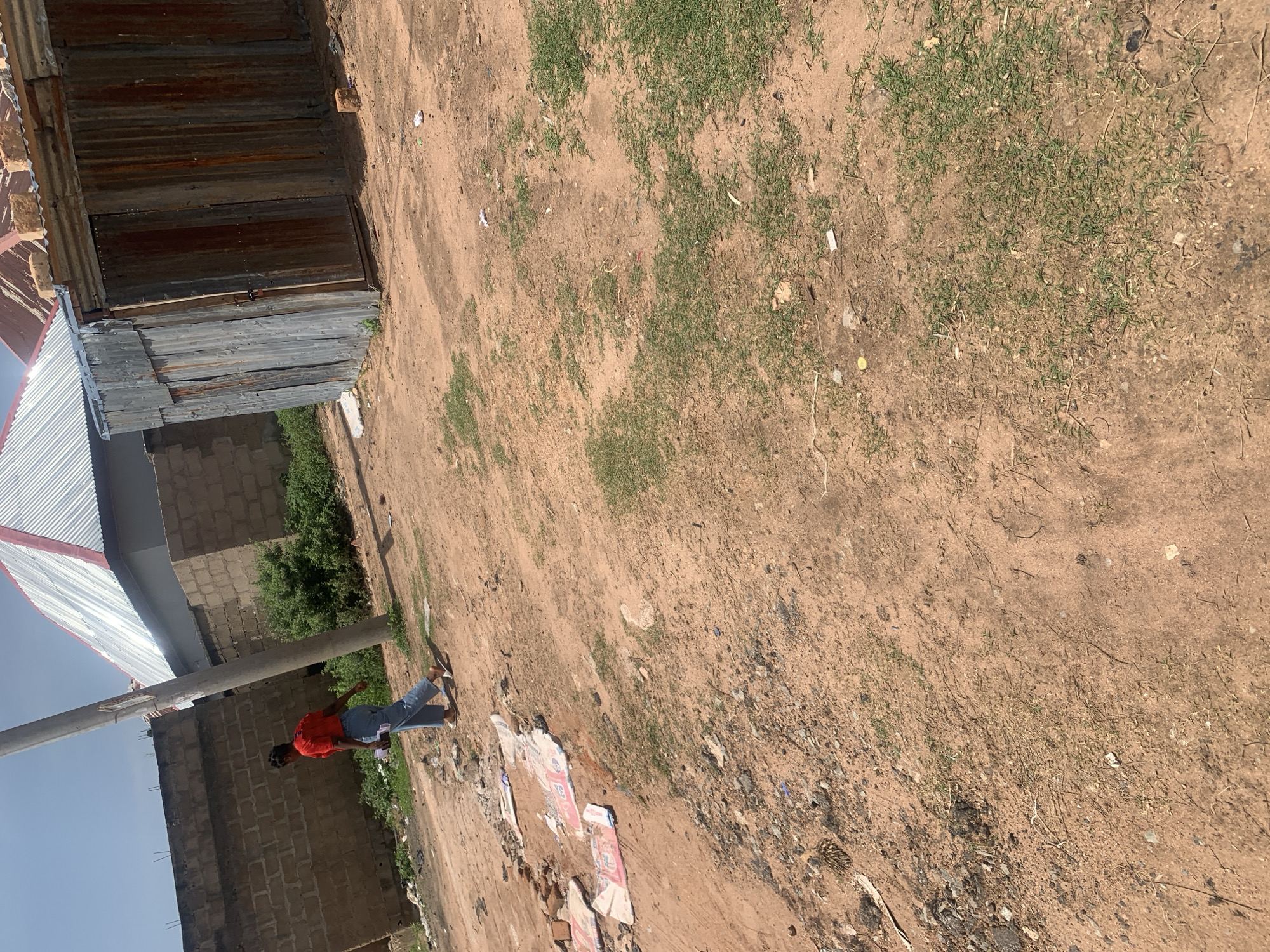 The image shows an outdoor scene in Jauro Yinu, Nigeria. A young woman wearing a red shirt and blue pants is walking on a dirt path. She is holding a white object in her left hand. To her left, there are some scattered pieces of material on the ground, possibly bags or wrappers. In the background, a brick wall and a corrugated metal structure are visible, along with a building with a silver metal roof. The ground is a mixture of dirt and patches of dry grass. A utility pole stands to the left of the woman.