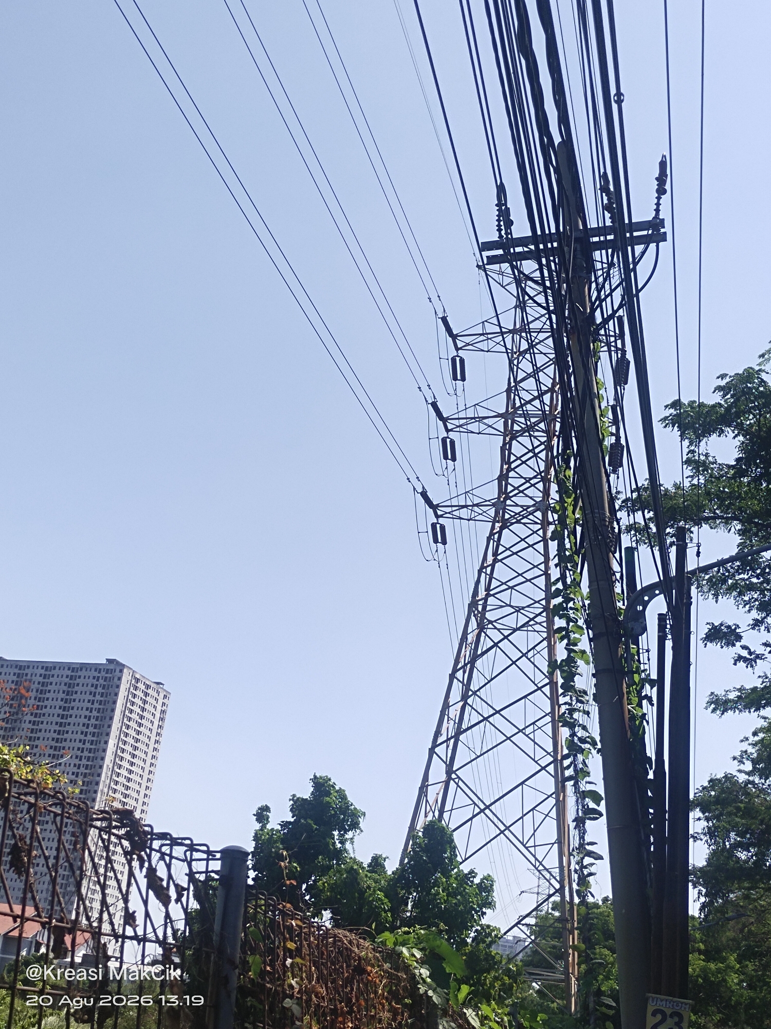 The image shows a view of a tall transmission tower with numerous power lines against a clear blue sky. In the background, a modern, multi-story building with a white facade and many windows is visible. A chain-link fence, partially covered in vines and foliage, runs across the foreground. Trees and other vegetation are scattered throughout the scene, with some vines growing up the sides of the transmission tower and poles. The overall scene suggests an urban or semi-urban environment in Surabaya, Indonesia, as indicated by the date and time stamp "@Kreasi MakCik 20 Agu 2026 13.19" which often includes location-specific information for social media posts or photo metadata.